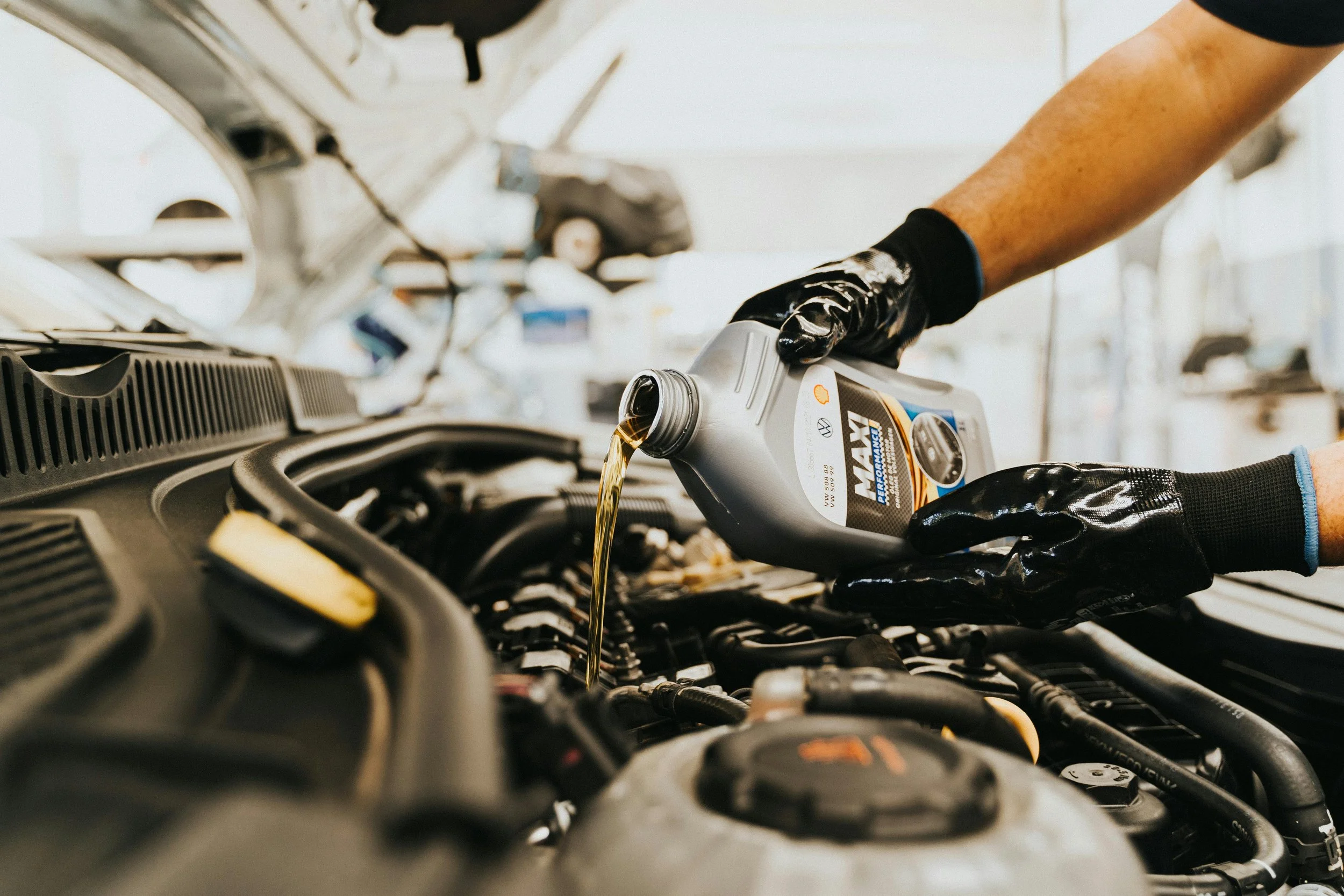 A mechanic wearing black gloves pouring motor oil from a gray bottle into a car engine.
