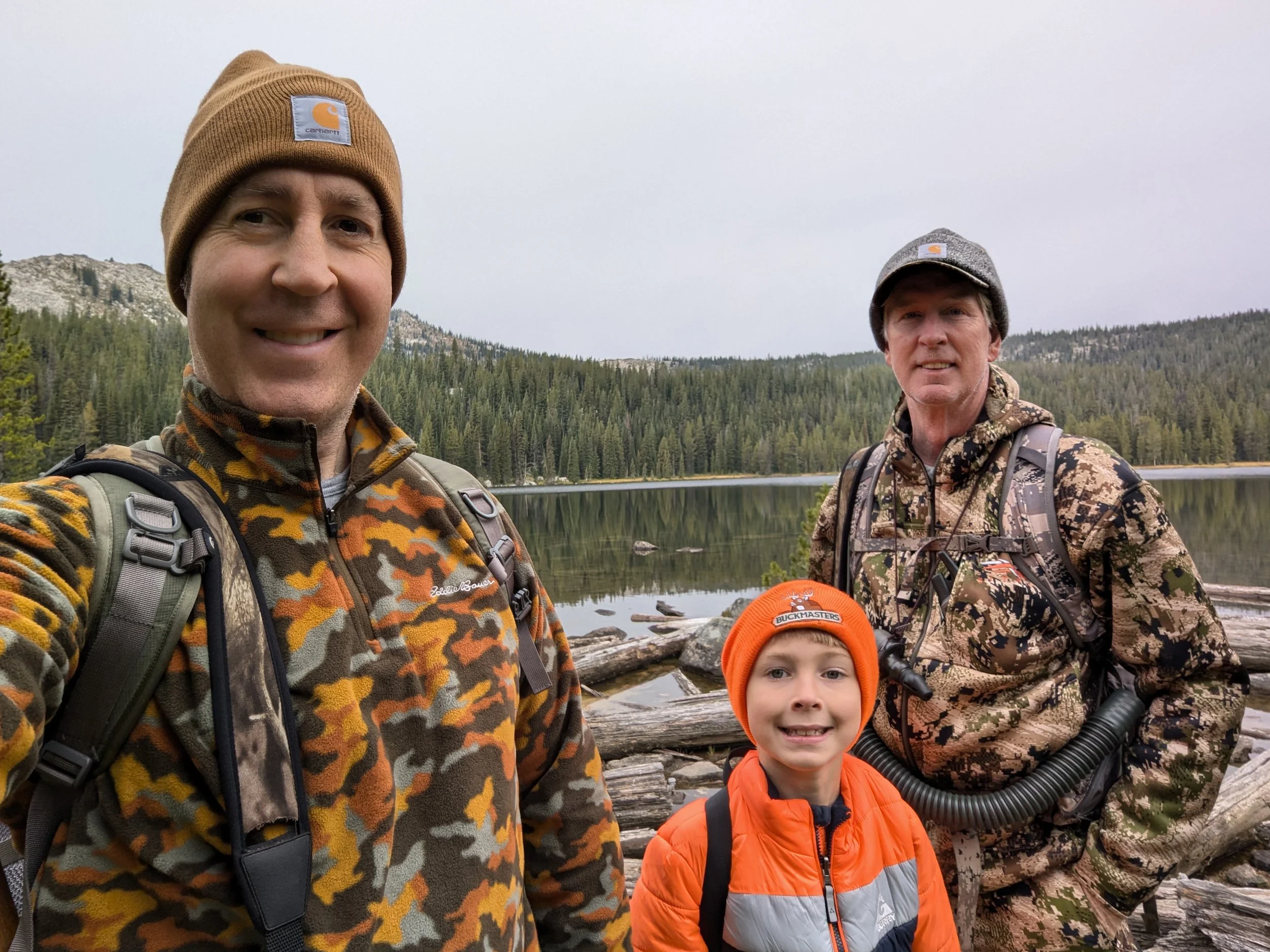 Three people, two adults and one child, taking a selfie outdoors near a lake surrounded by trees and mountains. They are dressed in camouflage and outdoor gear.