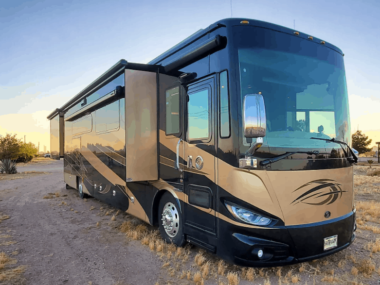 A large, modern motorhome with black and gold exterior paint parked on a dirt path in a desert landscape at sunset.