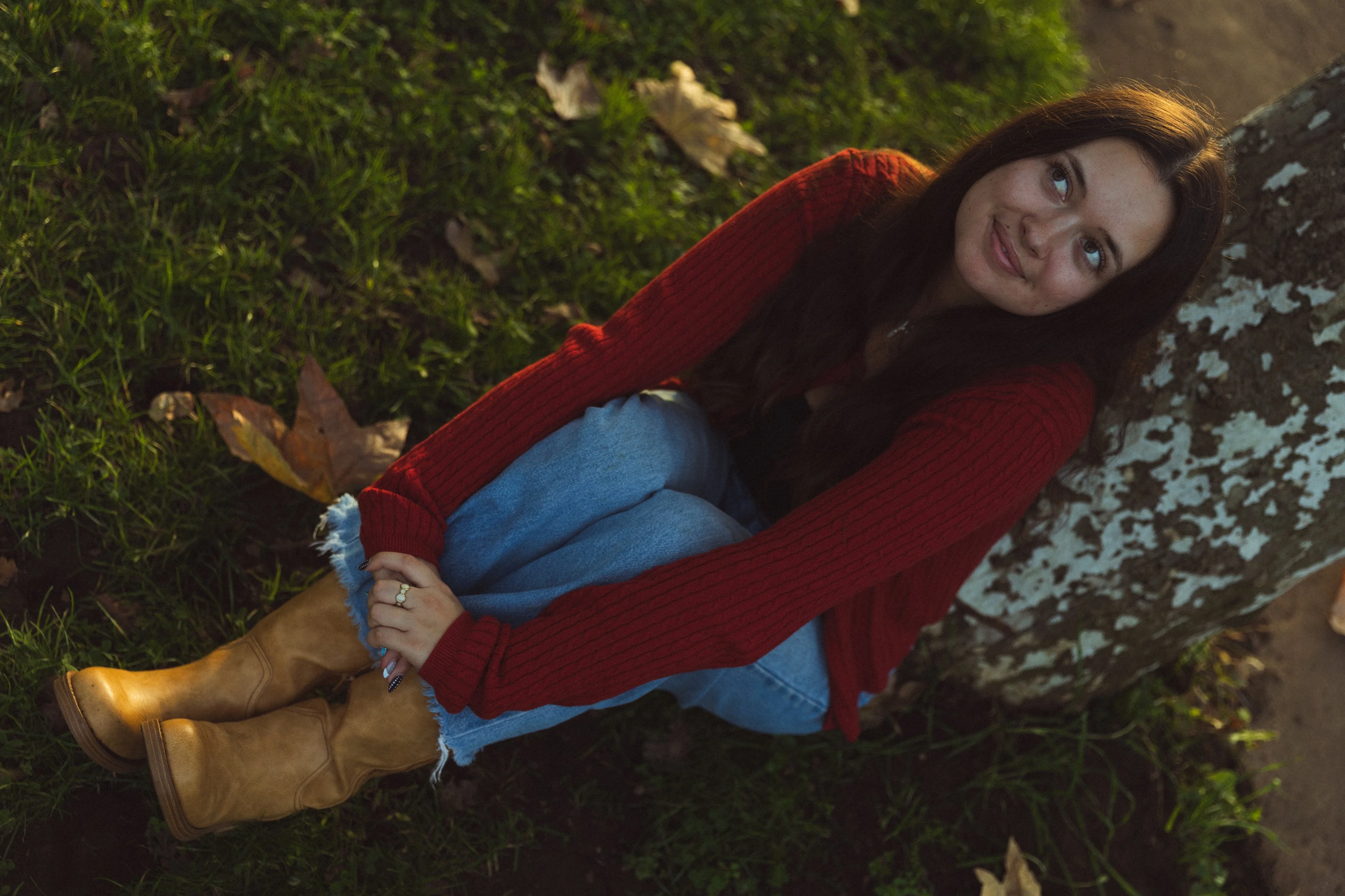A woman with long dark hair sitting on the grass next to a tree, wearing a red sweater, blue jeans, and tan boots, looking up and smiling.