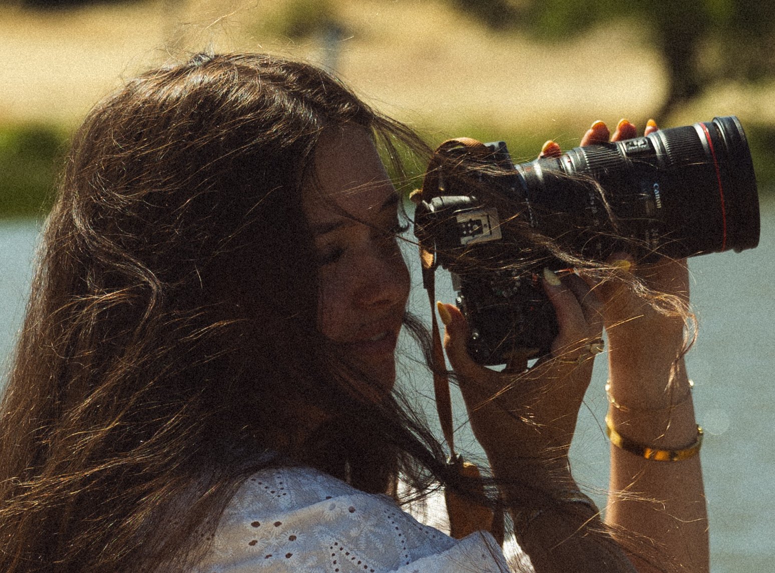A woman with long brown hair looking through a camera's viewfinder outdoors in sunlight.