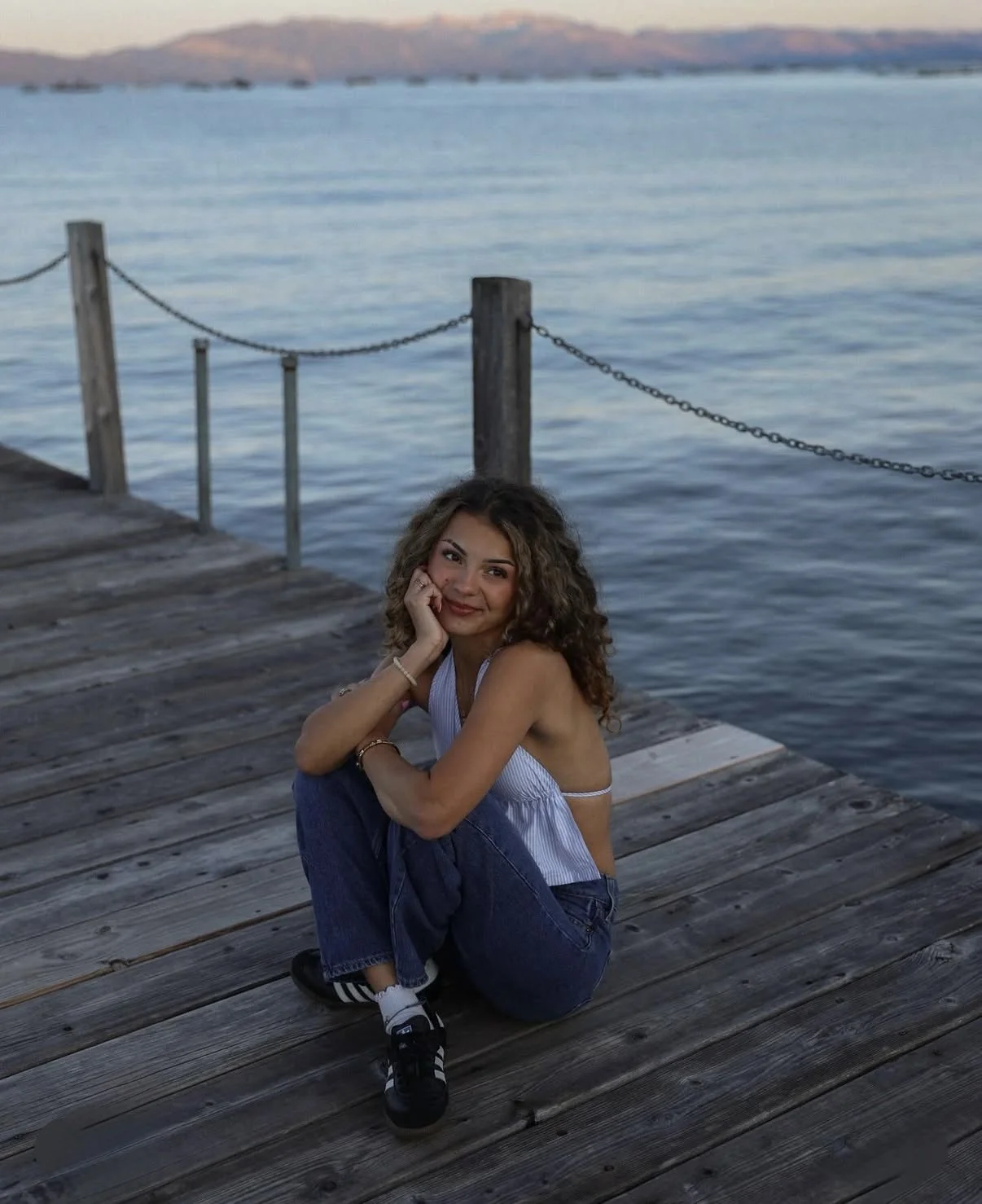 Young woman with curly hair sitting on a wooden dock near a body of water, smiling, wearing a white tank top, jeans, and sneakers, with a mountains in the background.