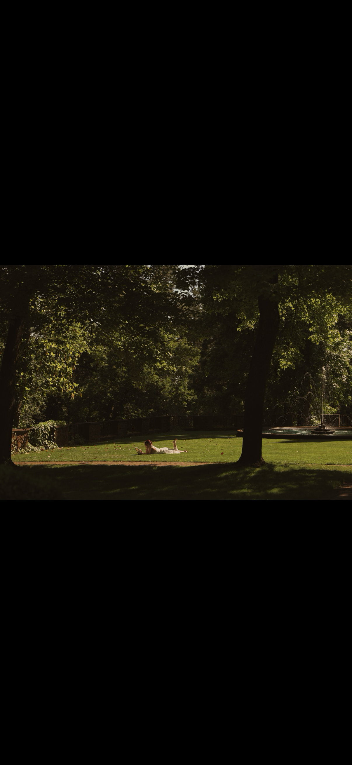 A person lying on a grassy park lawn reading a book, surrounded by large trees with green leaves, and a fountain in the background.