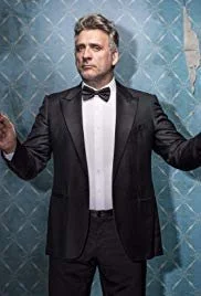 A man in a black tuxedo with a bow tie, standing with his arms extended, against a blue patterned background.