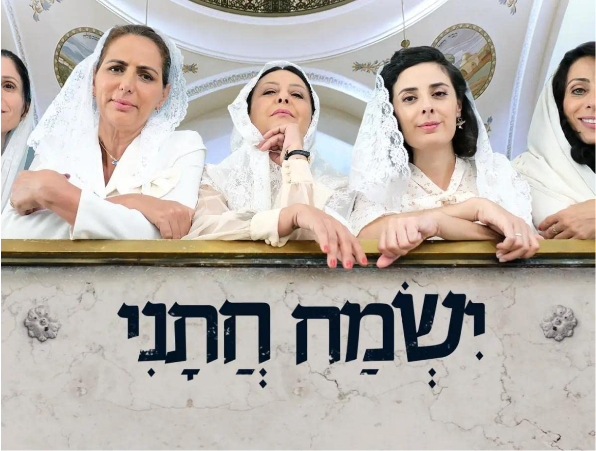 Four women in white attire with lace head coverings leaning on a marble balcony, inside a decorated building with Hebrew inscriptions on the front of the balcony.