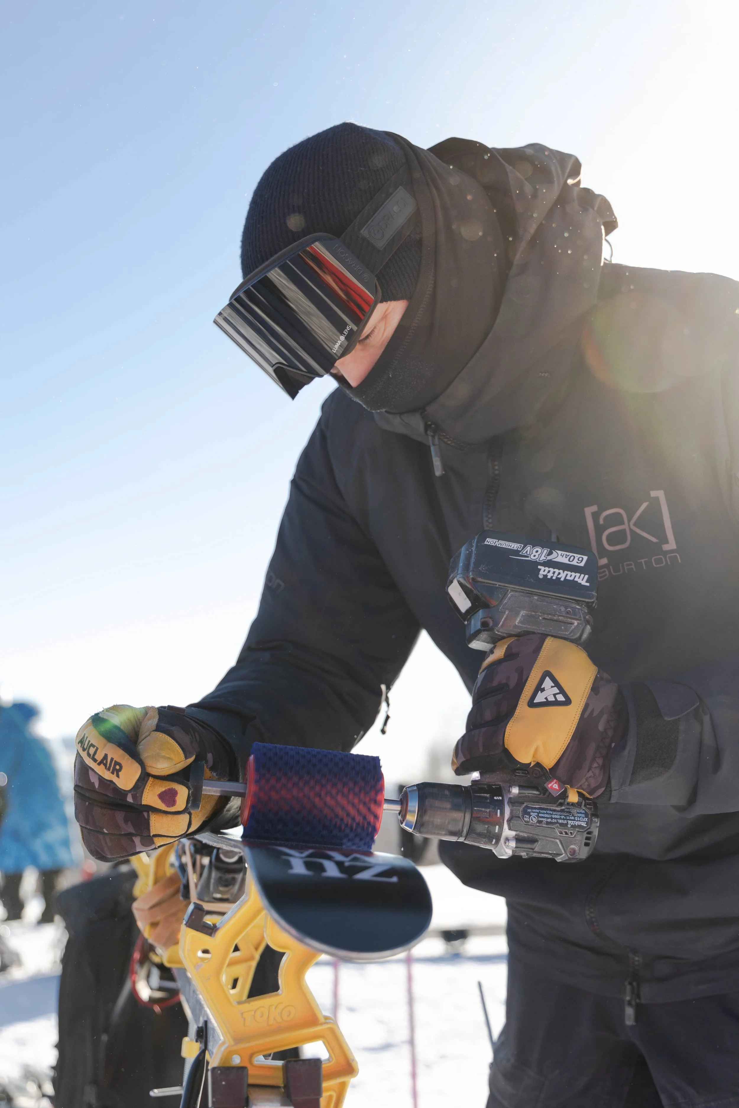 A person dressed in winter gear, including a beanie, sunglasses, and gloves, is working on a mountain bike in a snowy outdoor setting. The sun shines brightly in the sky.