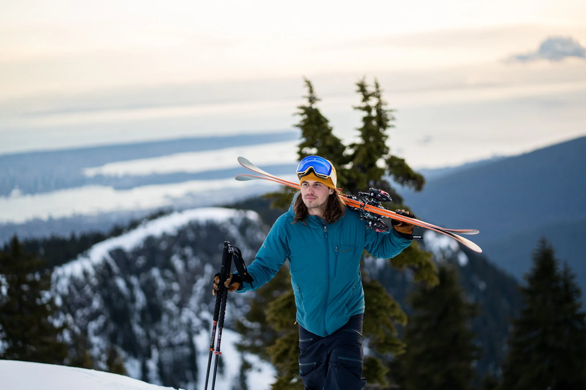 A man in a blue jacket holding skis and ski poles walking through snowy terrain with pine trees and mountains in the background during daytime.