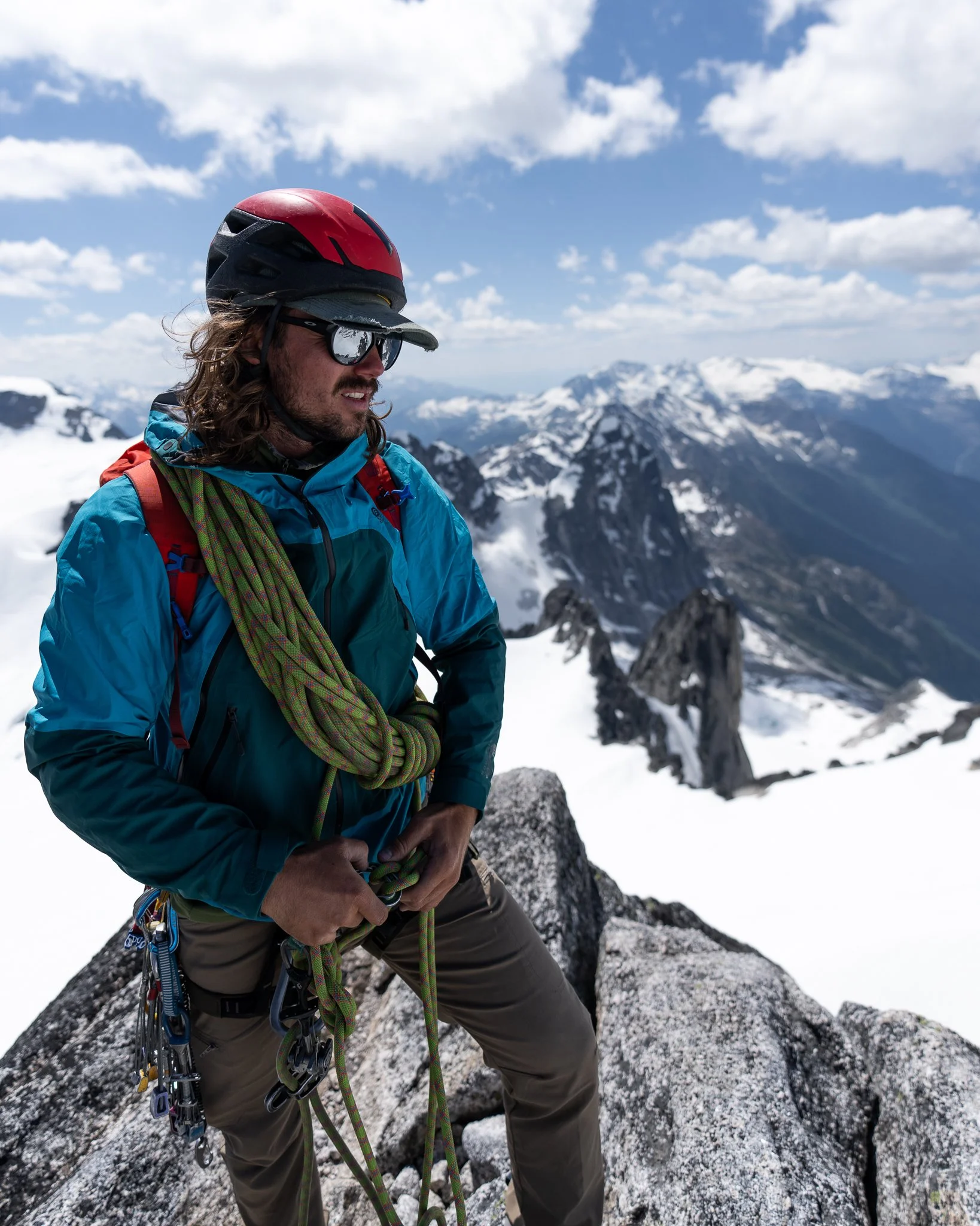 A man wearing a red helmet, sunglasses, blue jacket, and climbing gear standing on a snowy mountain peak with a mountain range and cloudy sky in the background.