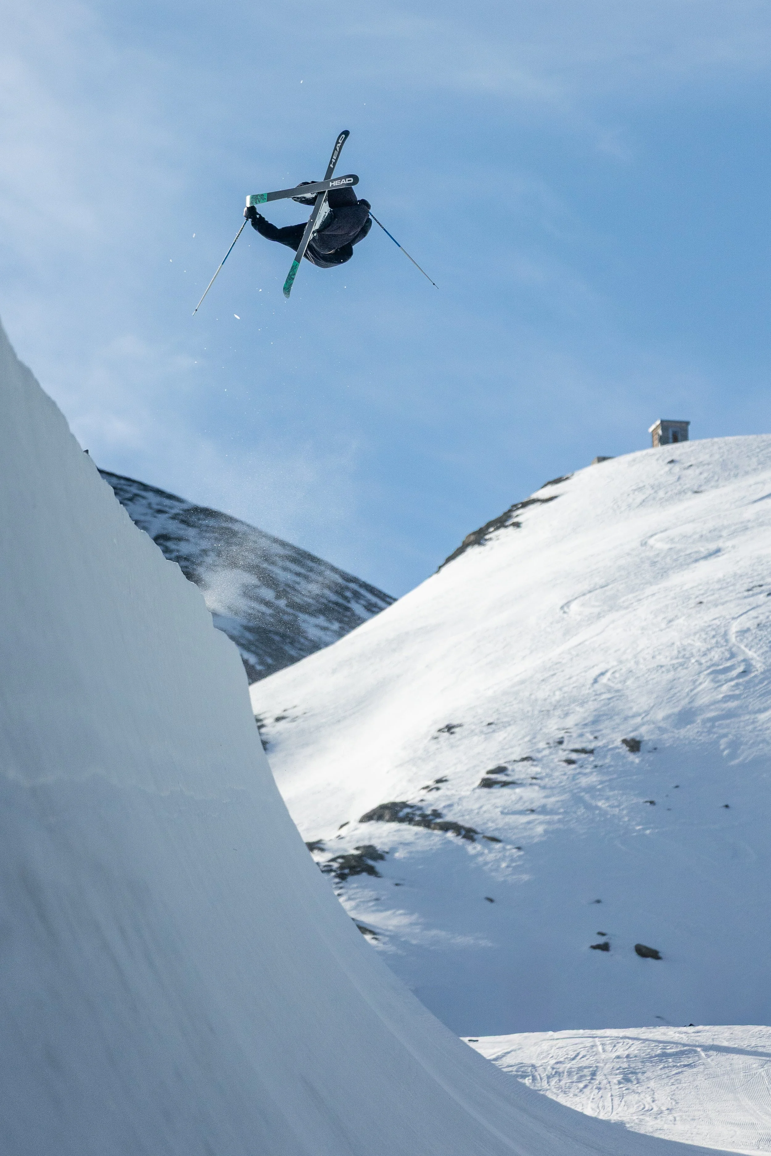 A skier in black gear executing a mid-air jump against a snowy mountain landscape and bright blue sky.
