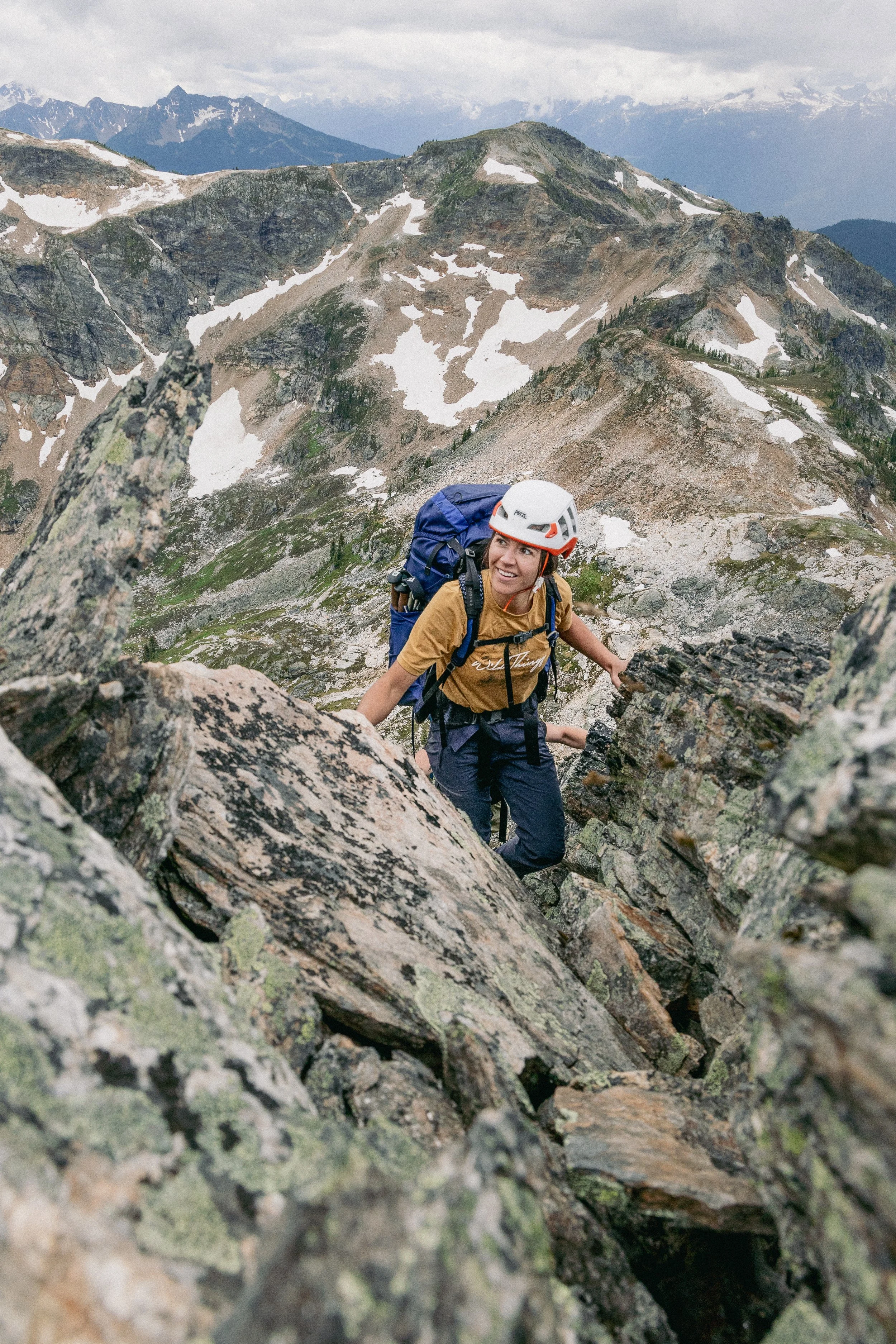A woman climbing a rocky mountain trail with a blue backpack and a white helmet, surrounded by mountain peaks and patches of snow.