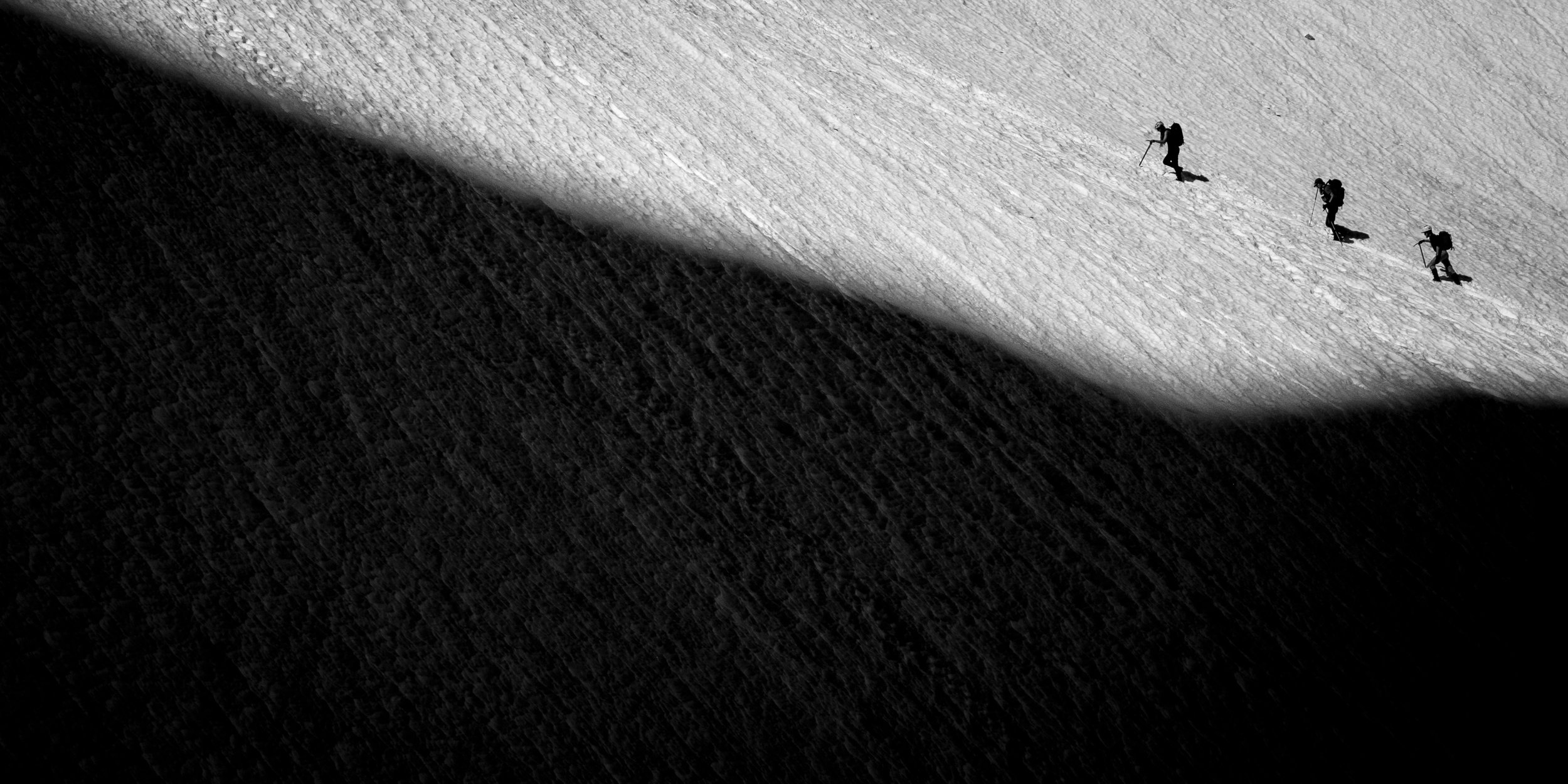 Silhouettes of three hikers with backpacks walking up a snow-covered mountain slope, with a sharp shadow dividing the snowy and rocky terrain in black and white.