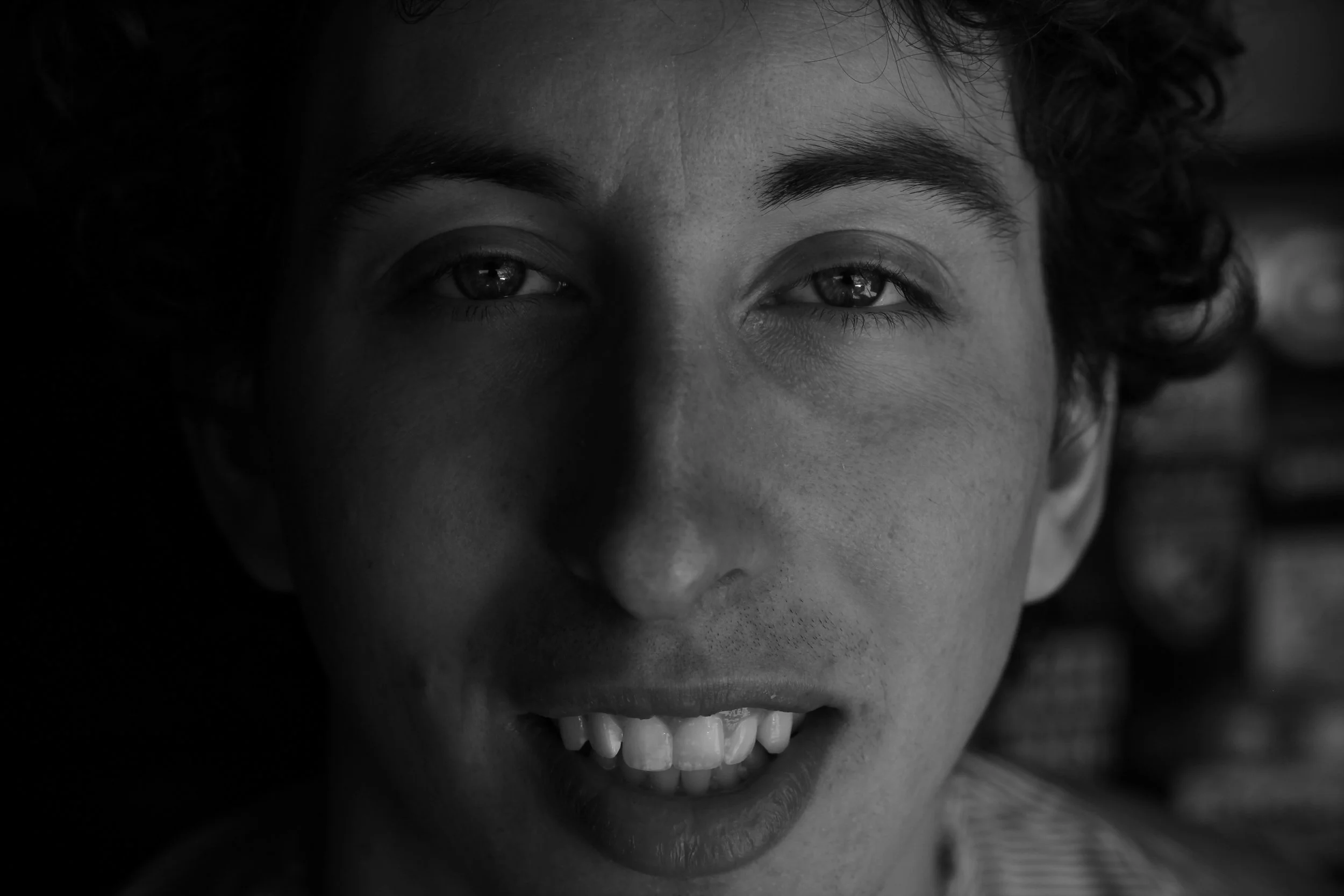 Black and white close-up of a person smiling, showing their teeth, with curly hair and expressive eyes.
