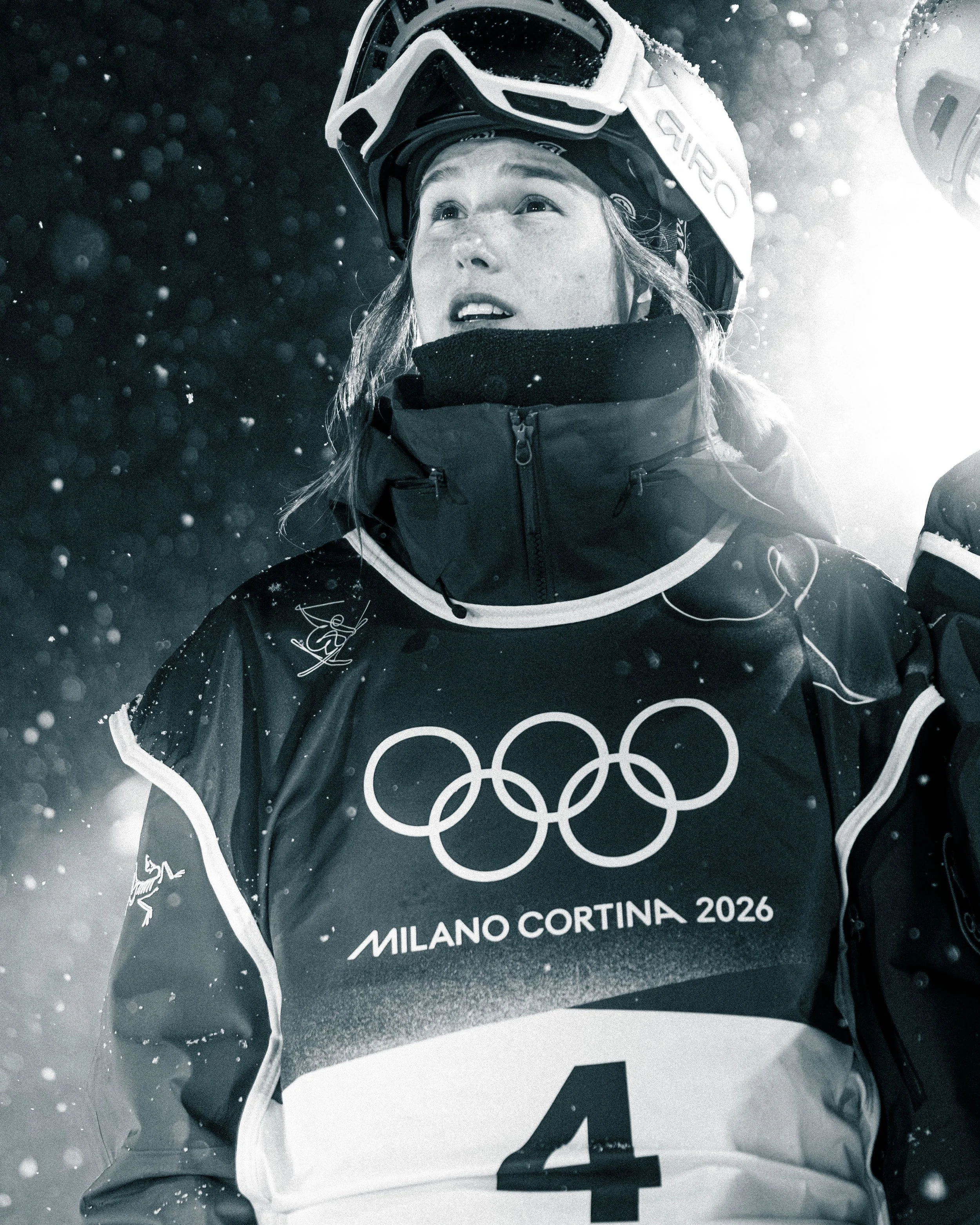 Close-up of a person wearing a helmet with a ski mask and winter gear, looking upwards with snow falling around, indicating a winter sports setting at the Milano Cortina 2026 Olympics.
