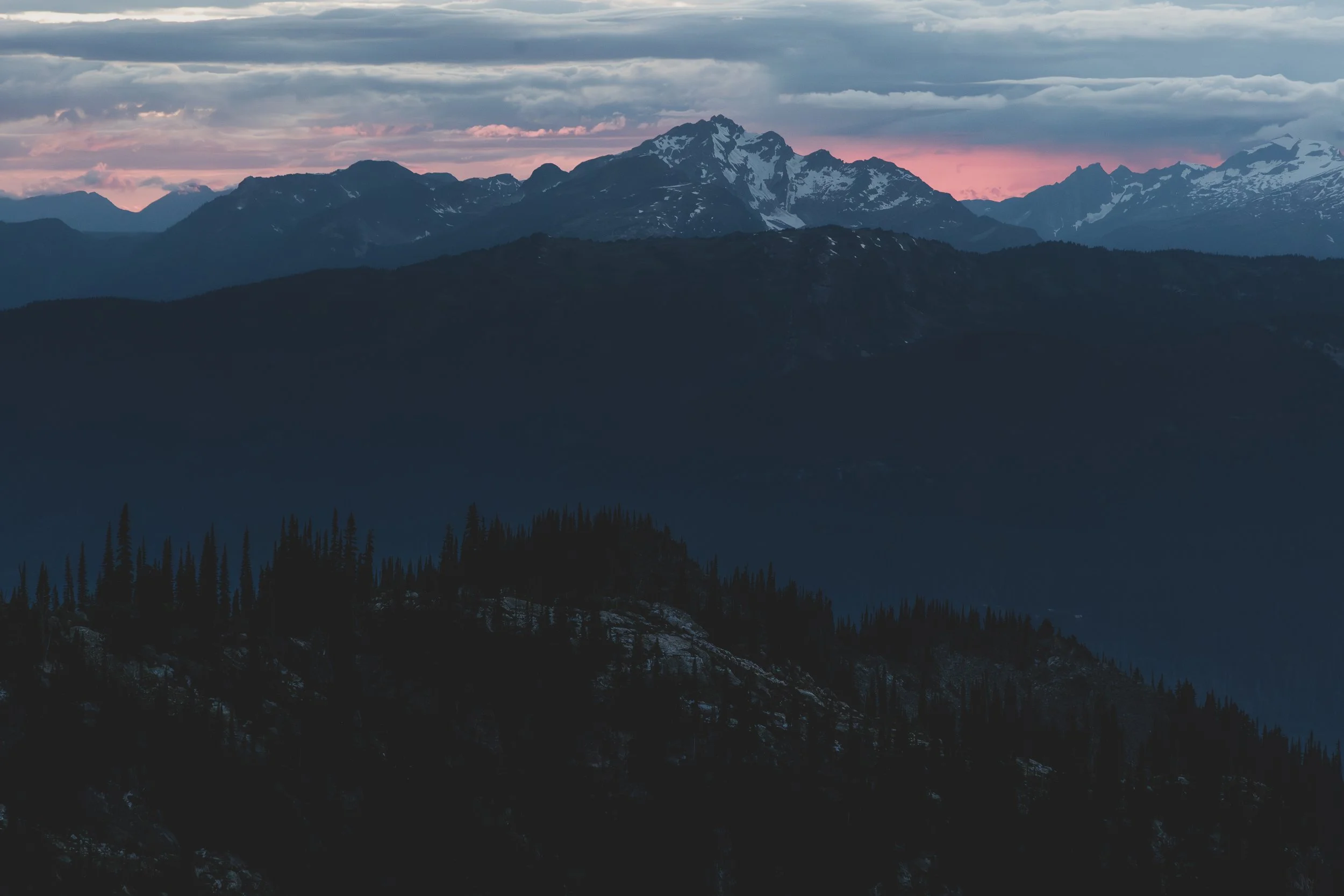 Scenic view of snow-capped mountains under a cloudy sky during sunset, with a forested hillside in the foreground.