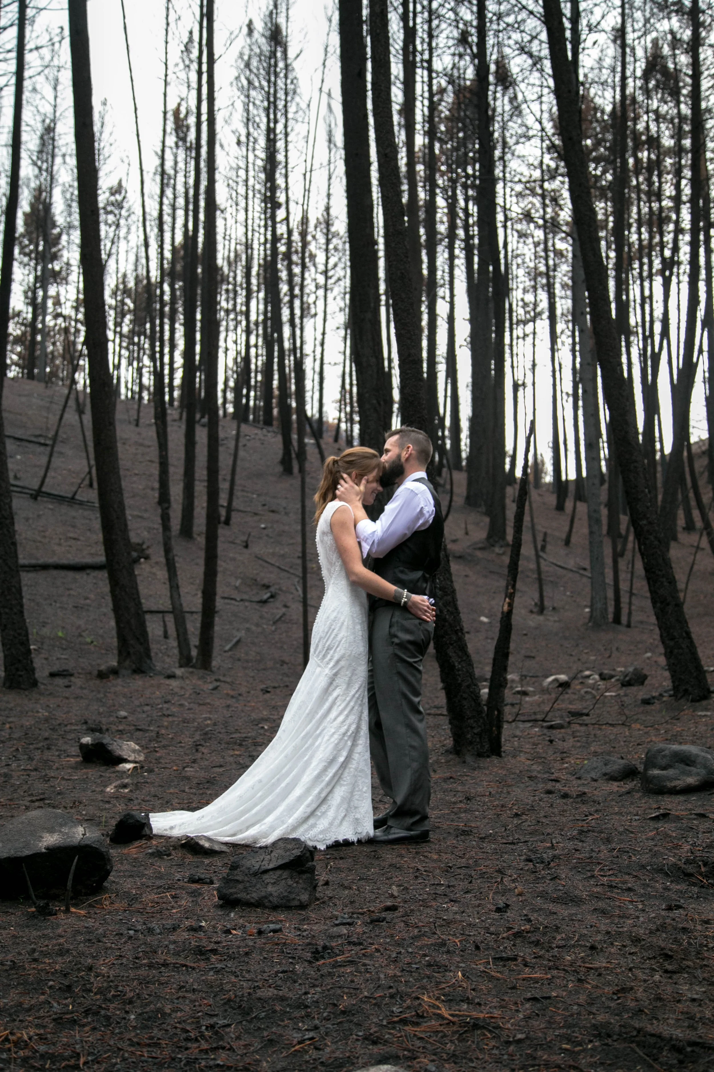 A couple dressed in wedding attire standing close together in a charred forest, with the woman wearing a white wedding dress and the man in a black vest and gray pants.