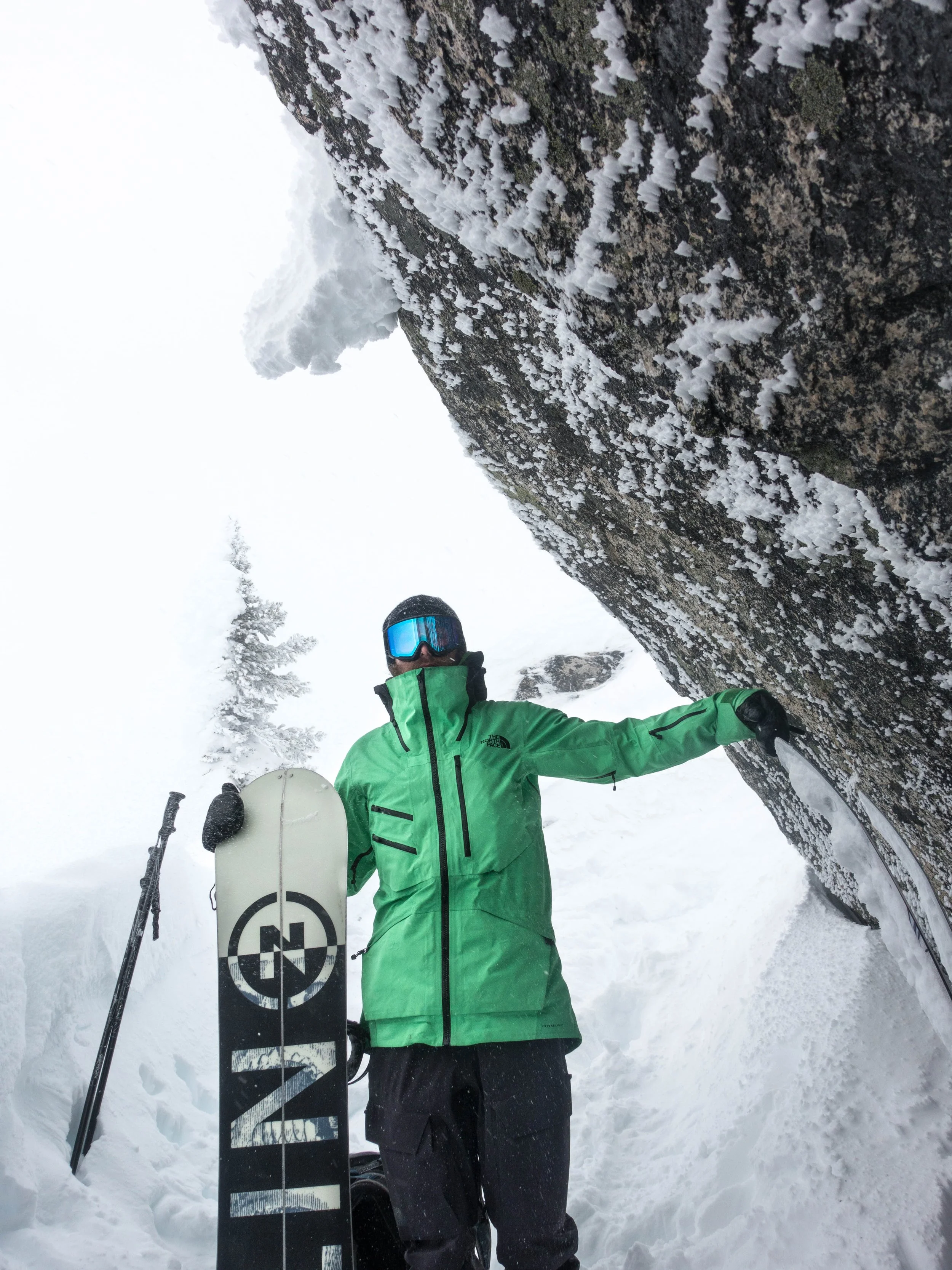 A person wearing a bright green jacket and black pants, holding a snowboard, standing in the snow near a large snow-covered tree in a snowy landscape, with ski poles leaning nearby. The person is also wearing goggles and gloves.