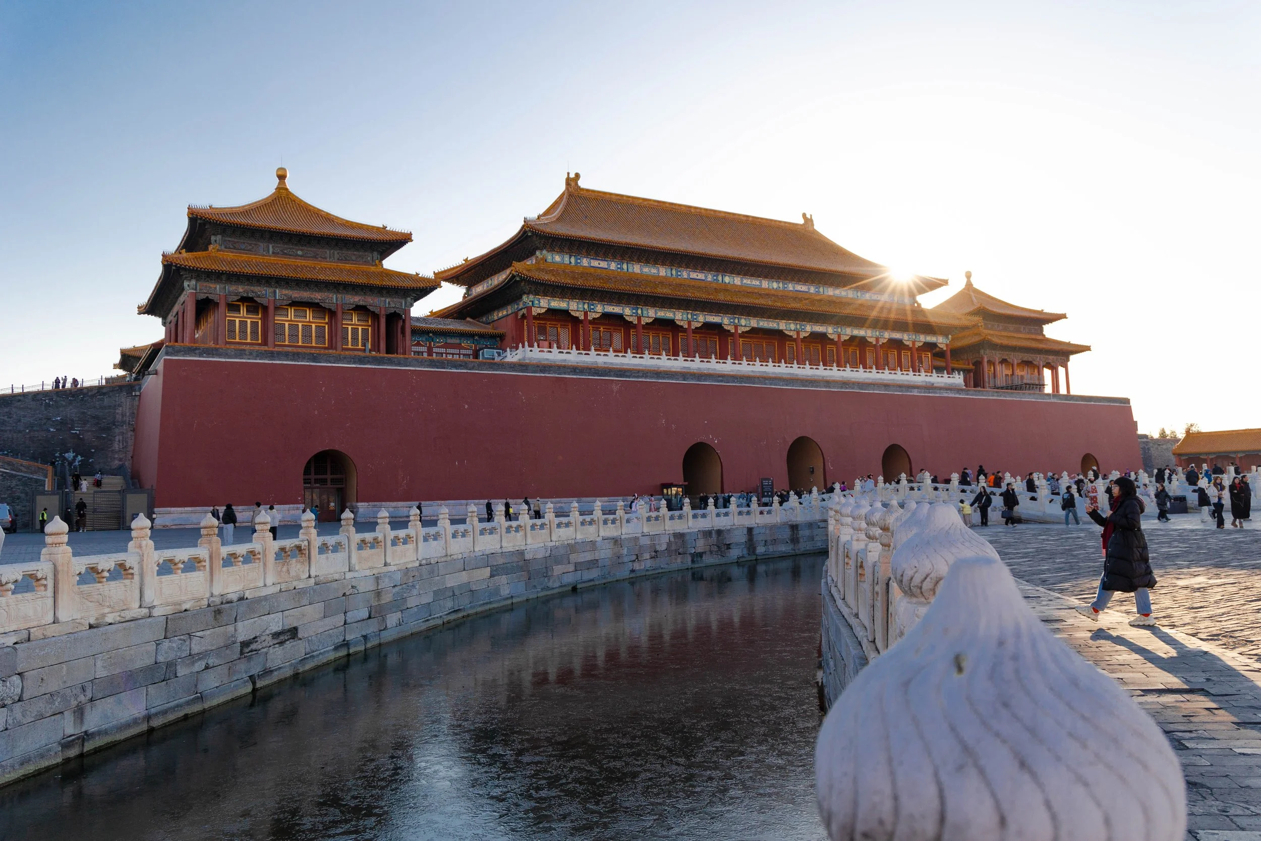 Traditional Chinese palace with red walls and golden roofs, situated above a waterway with stone dividing walls and a white marble balustrade, people walking around in the foreground, during sunset.