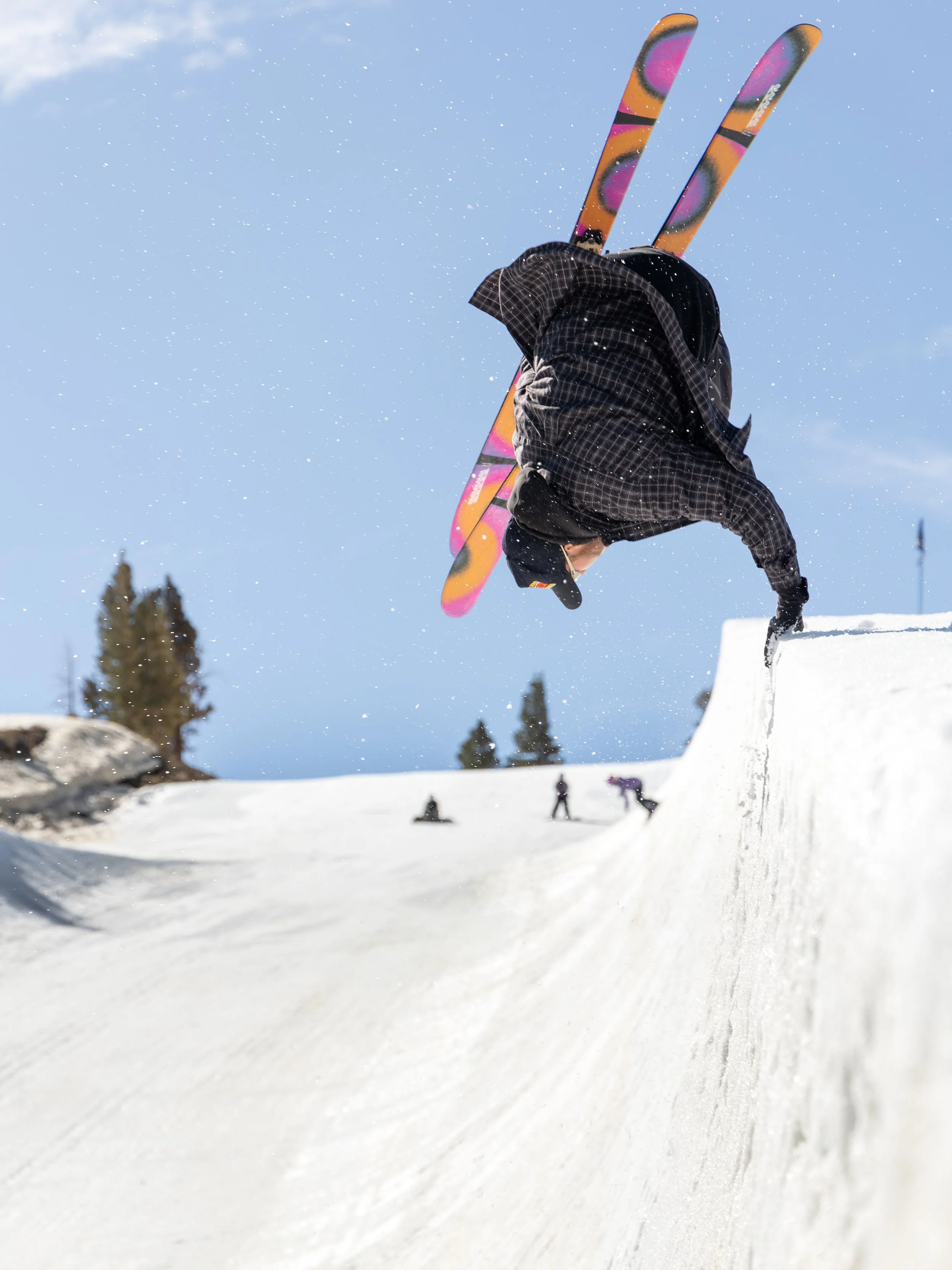 Skier in black and checkered jacket performing a backflip on a snow-covered ski slope with other skiers in the background.