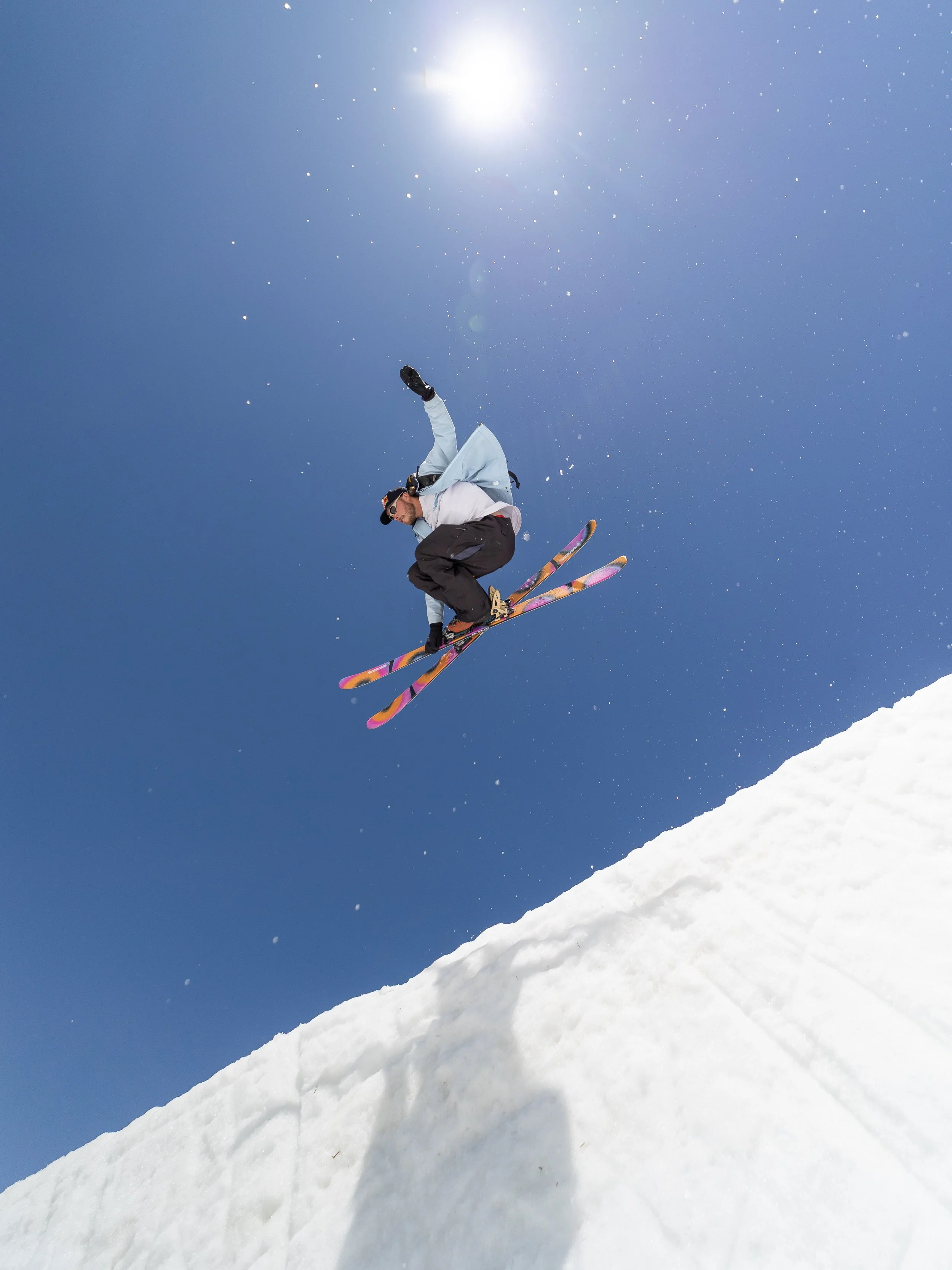 A skier jumping in mid-air on a snowy slope under a bright sunny sky with a few clouds.