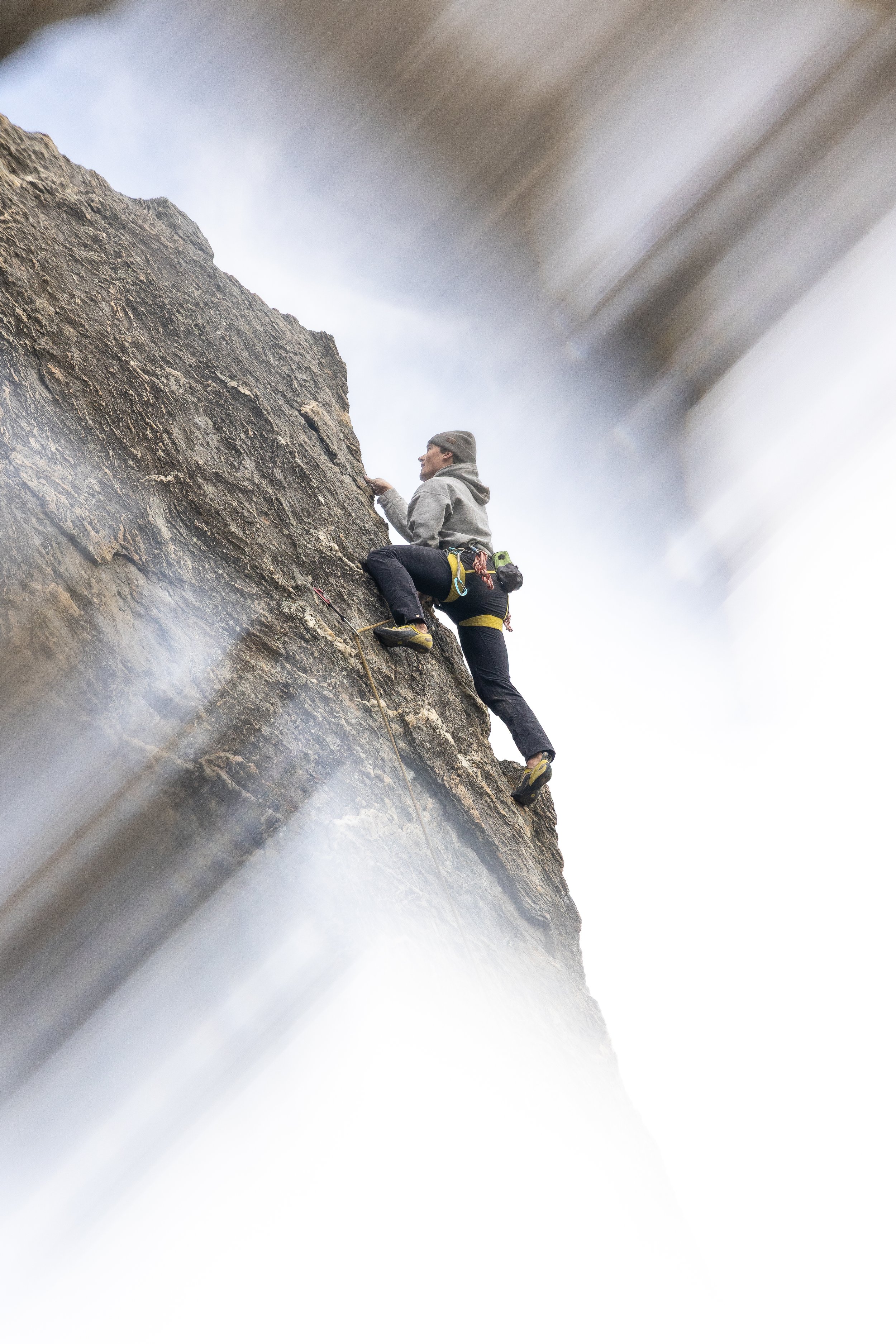 A person rock climbing, wearing a gray hoodie, black pants, climbing shoes, and a beanie, ascending a steep rock face using climbing gear.
