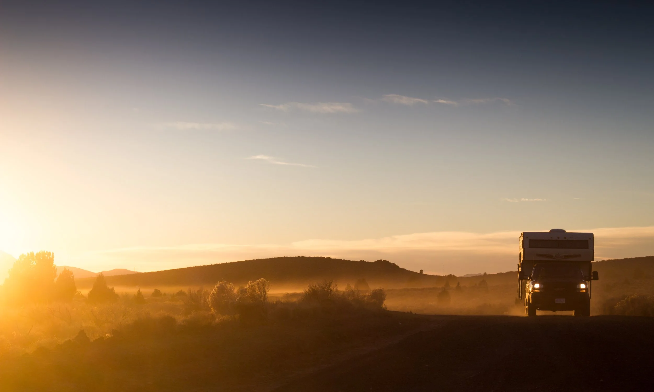 A black camper van driving on a rural road during sunset with hills and trees in the background and a partly cloudy sky.