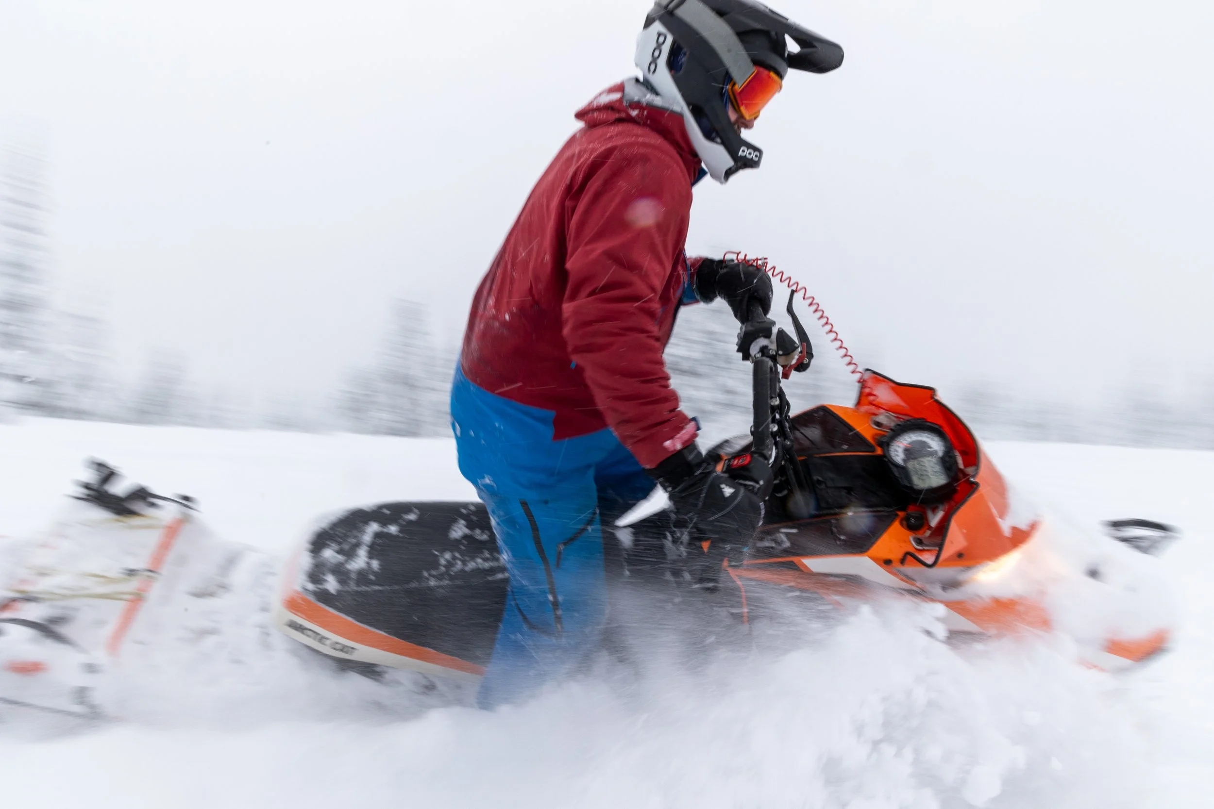 Person riding an orange snowmobile in snowy landscape, wearing a helmet and winter gear.