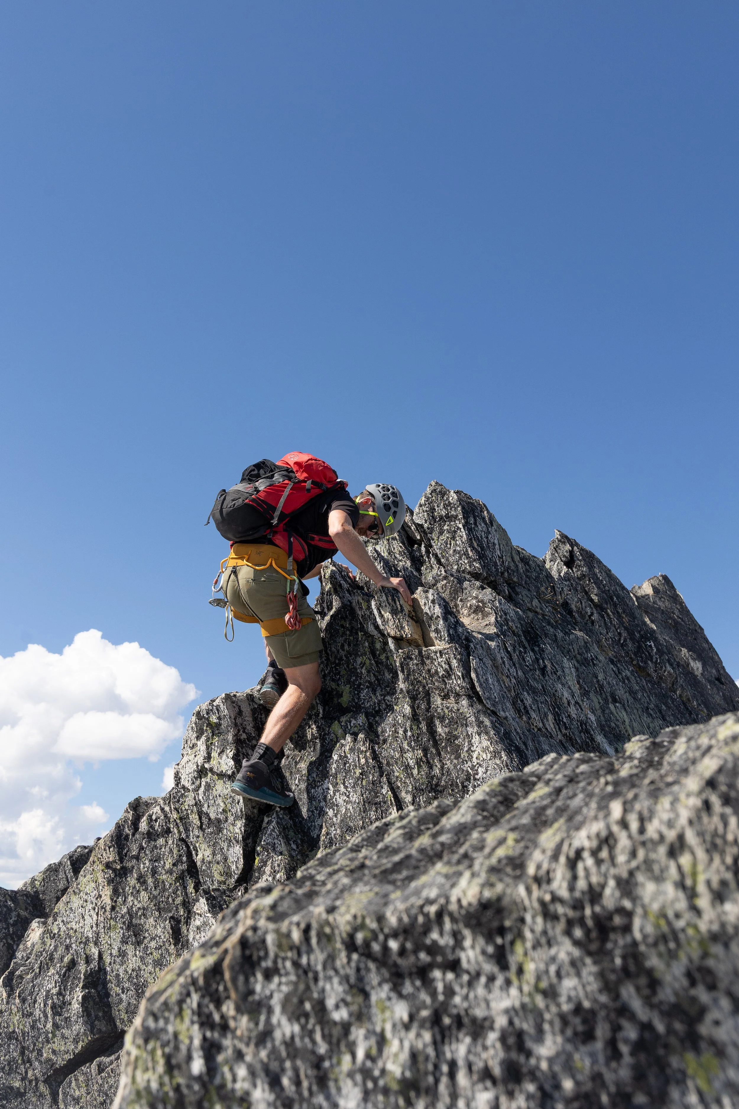 A person with a helmet, backpack, and hiking gear climbing on large rocks in a mountainous area.