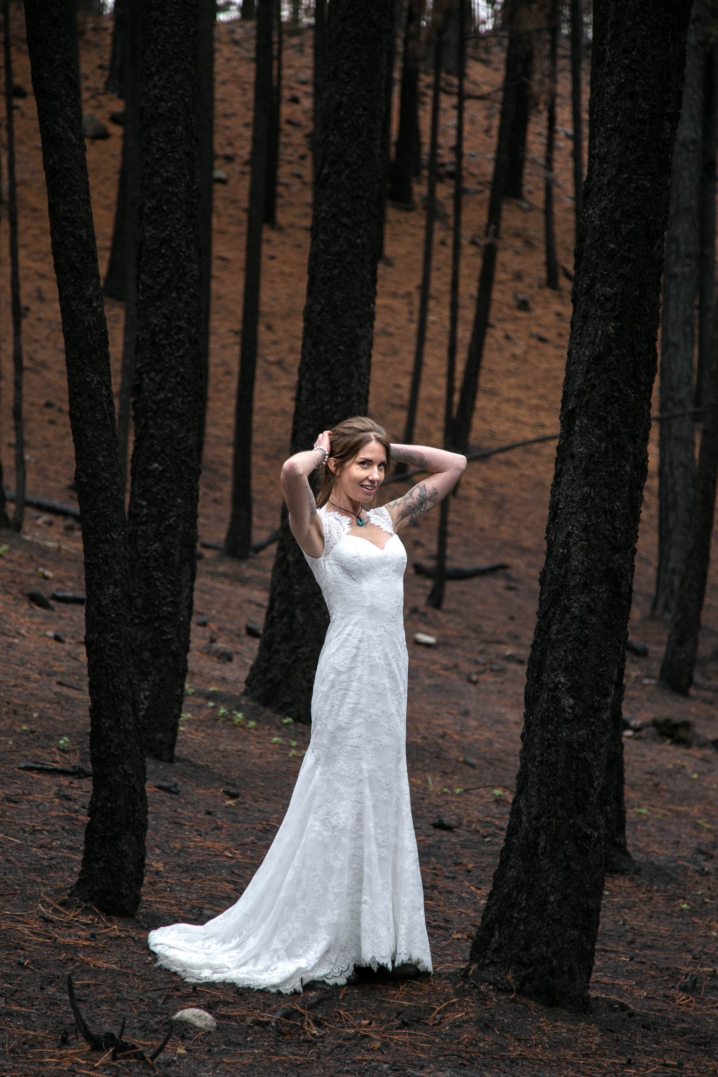 A woman in a white wedding dress standing in a forest with burnt trees, smiling and adjusting her hair.