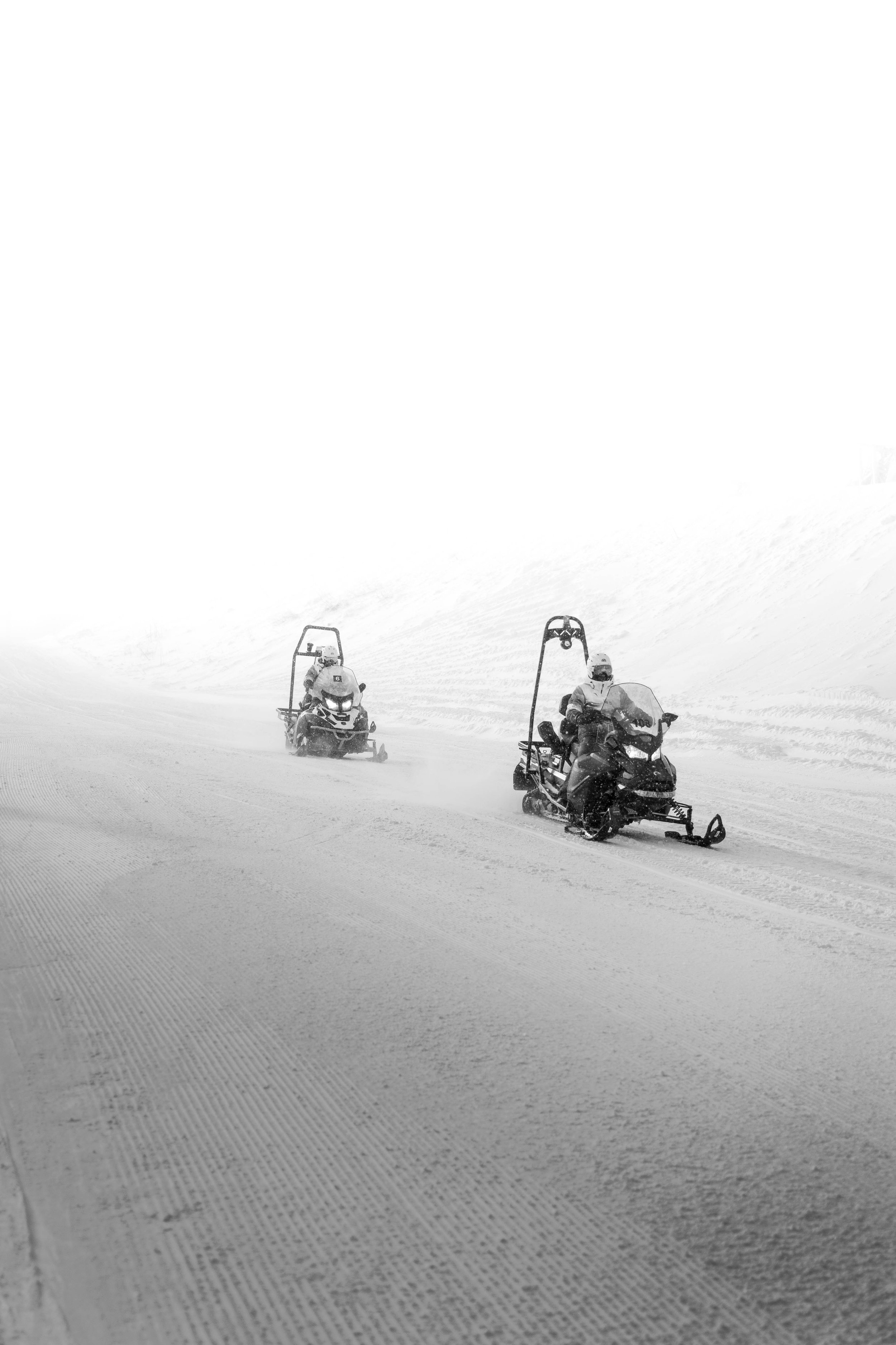 Two snowmobiles driven by people in winter gear on a snowy landscape with snow-covered ground and a foggy or snowy background.