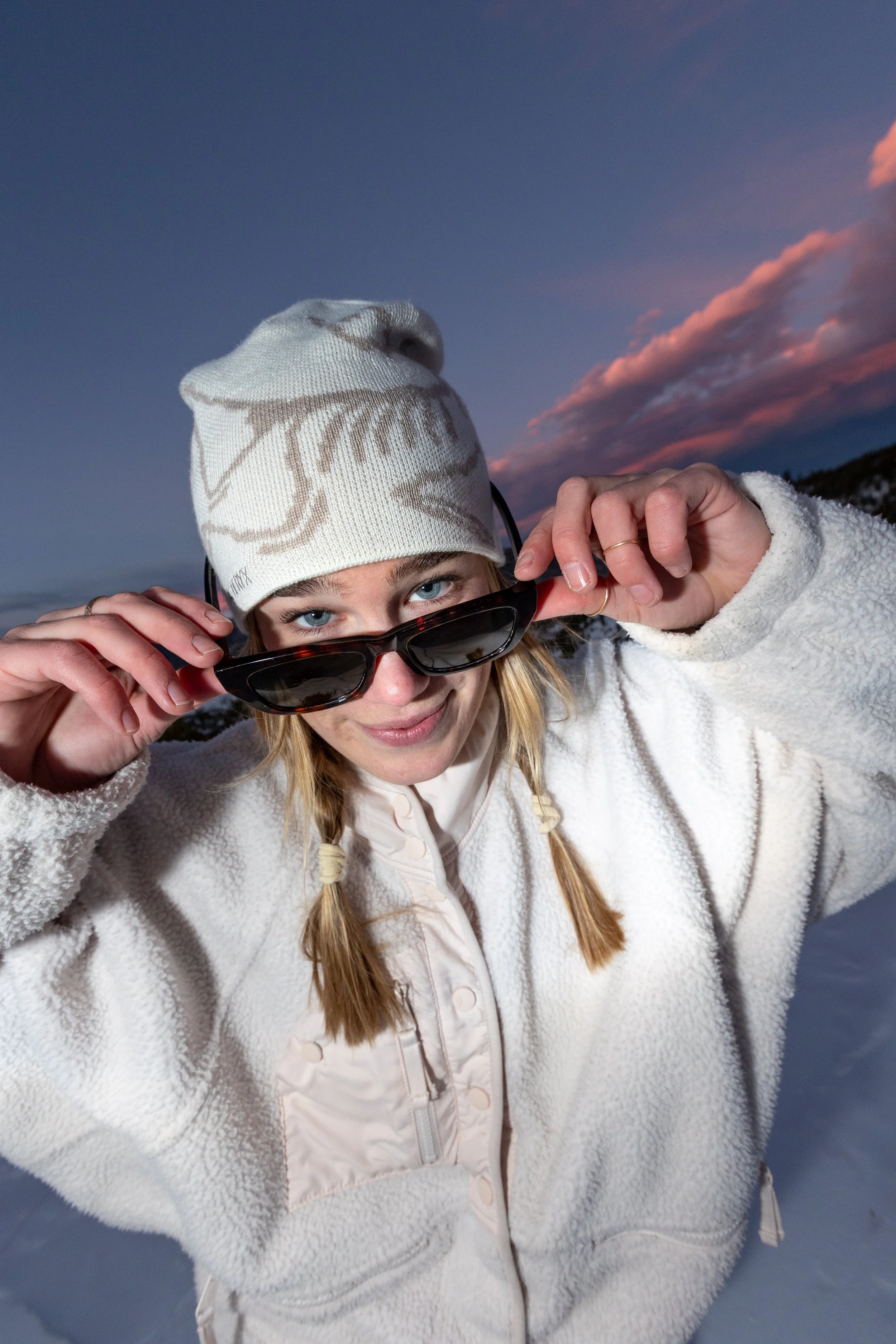 A young woman with blue eyes, wearing a white fleece jacket, a white winter hat with patterns, and black sunglasses, adjusts her sunglasses outdoors during dusk with a colorful sunset sky and snow-covered ground in the background.