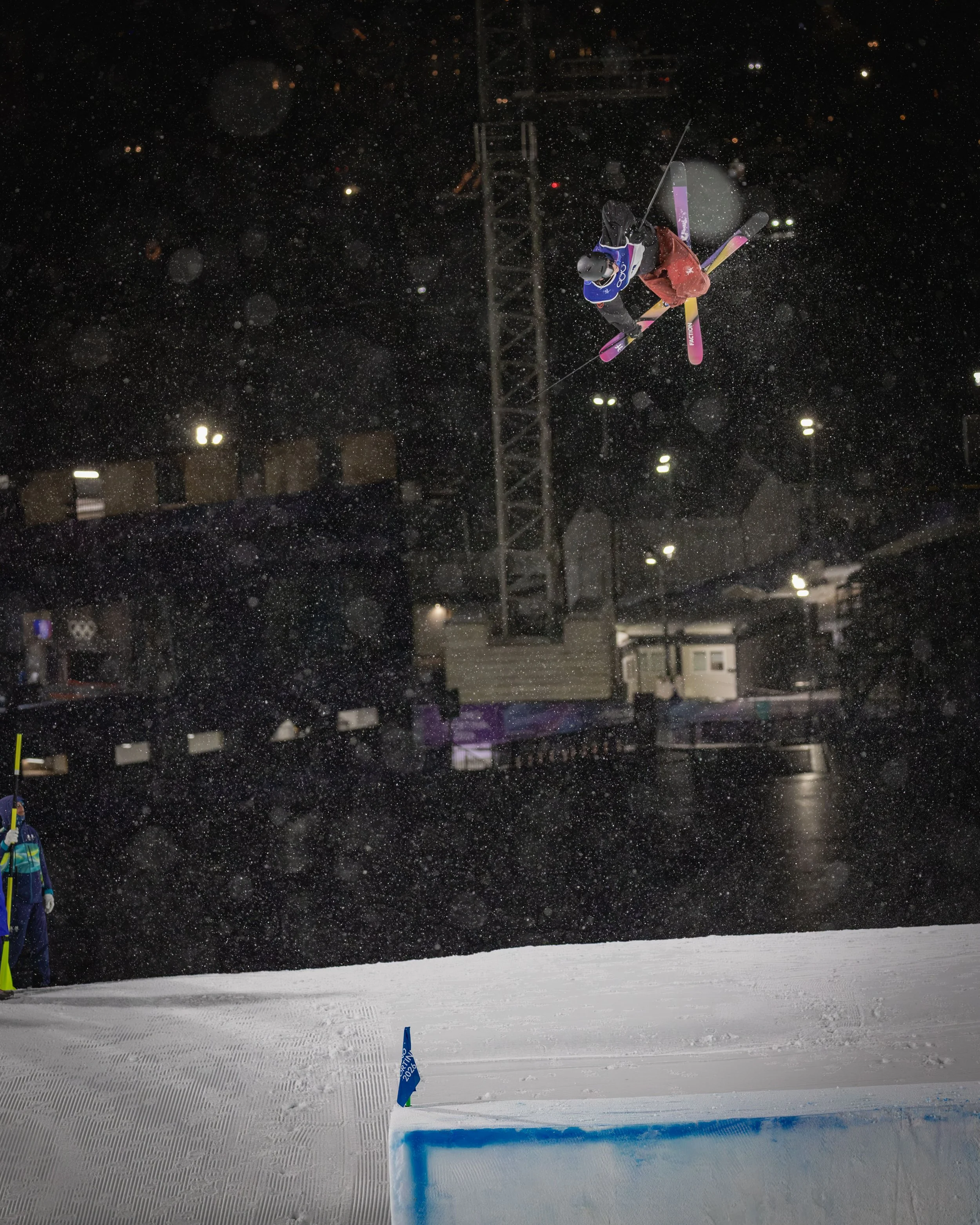 Nighttime ski jump competition with a skier mid-air above snow and a snow feature with a blue edge in the foreground. Snow is falling, illuminated by bright lights.