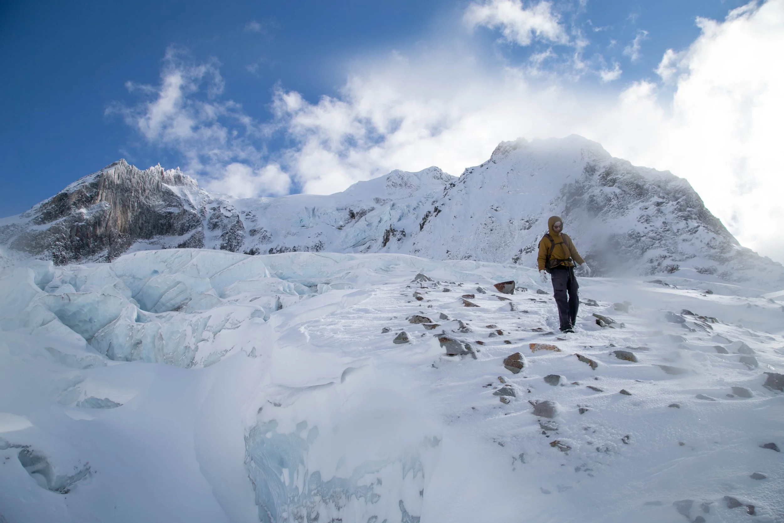 A person in winter gear, including a yellow jacket, black pants, and a hat with goggles, walking on snow and ice in a mountainous, glacial landscape with snow-covered peaks and a partly cloudy sky.
