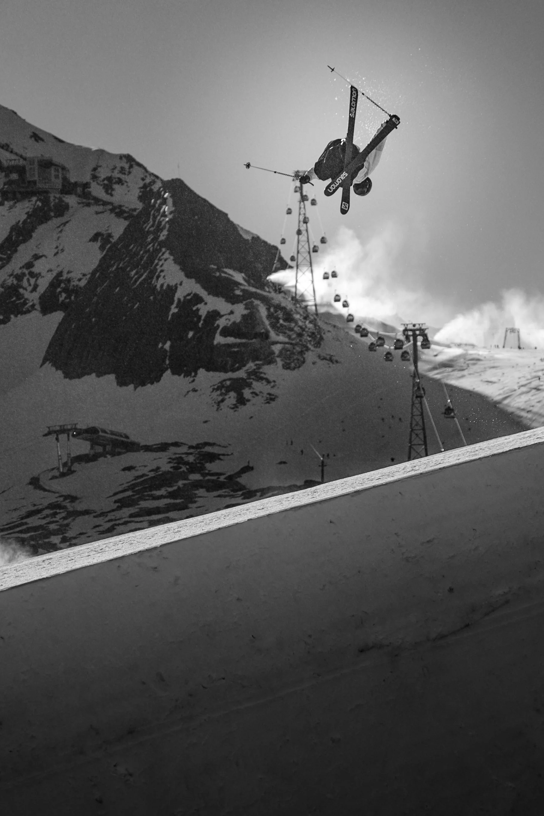 A skier performing a jump in mid-air at a snowy mountain ski resort with ski lifts and a mountain peak in the background.