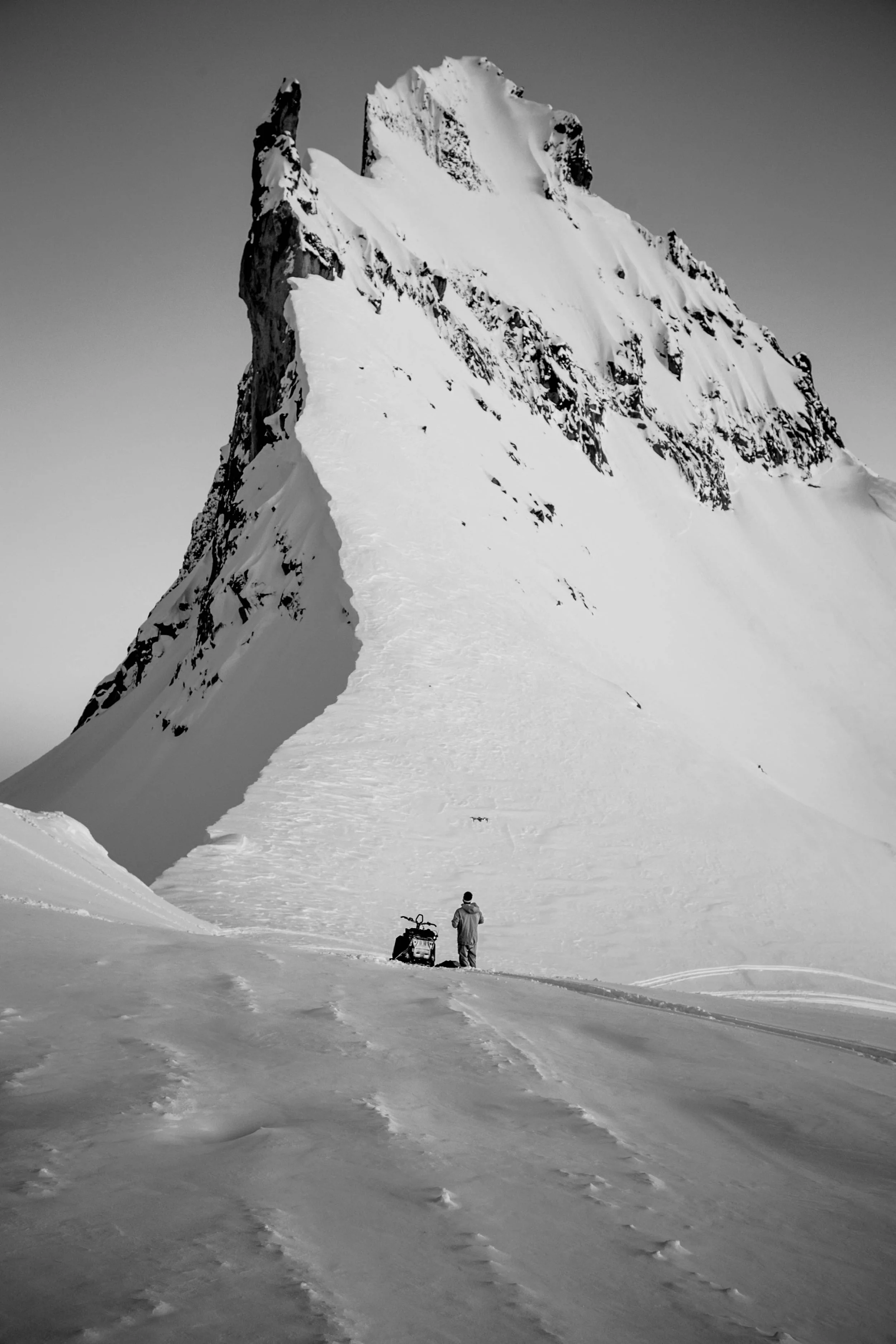 A person standing on snowy terrain with a snowmobile, gazing at a tall, snow-covered mountain in the distance.