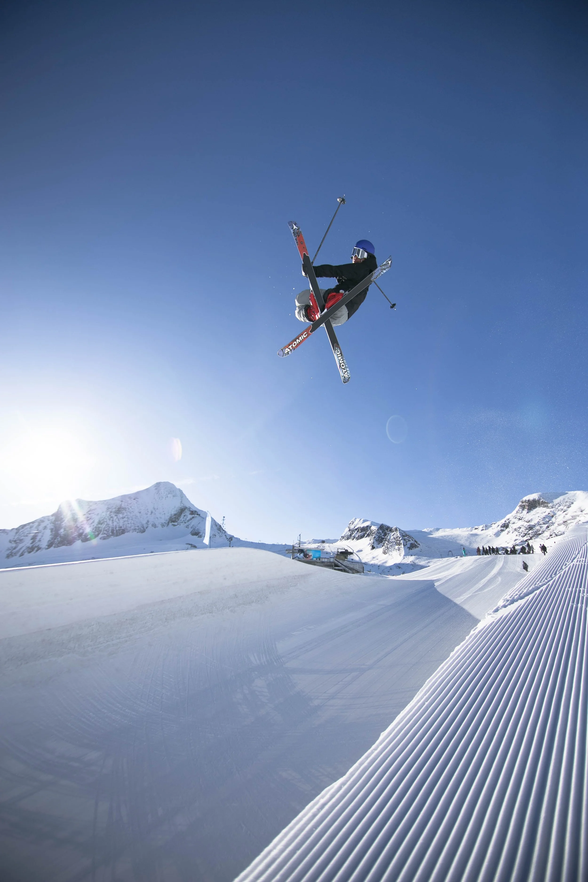 Skier airborne over snow-covered ski slope with mountains in the background under a clear blue sky.