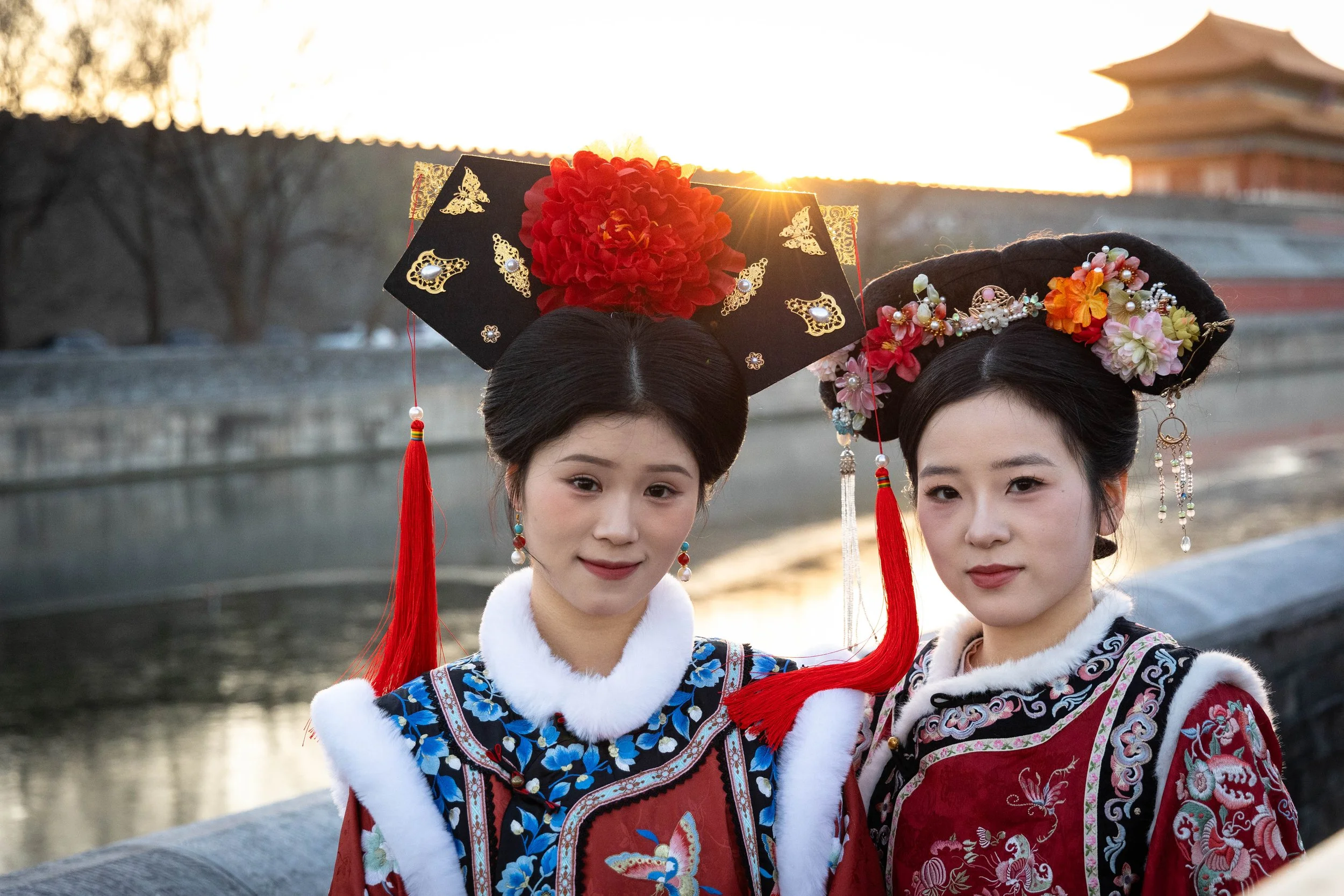 Two women dressed in traditional Chinese opera costumes with ornate headdresses standing outdoors near a river at sunset.