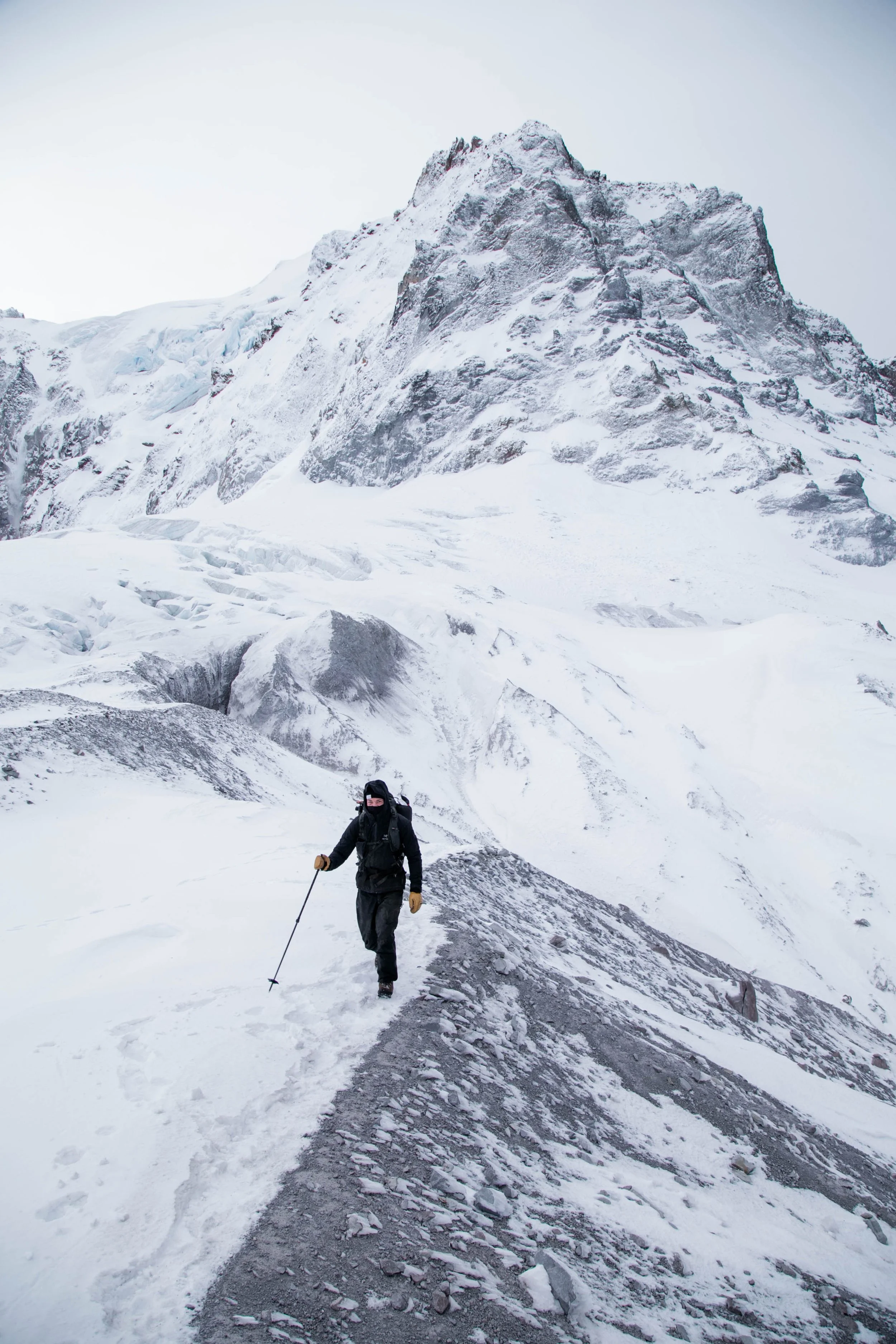 A climber wearing a black outfit, hat, gloves, and goggles trekking across a snow-covered mountain trail with a hiking pole, in a rugged, snowy mountain landscape.