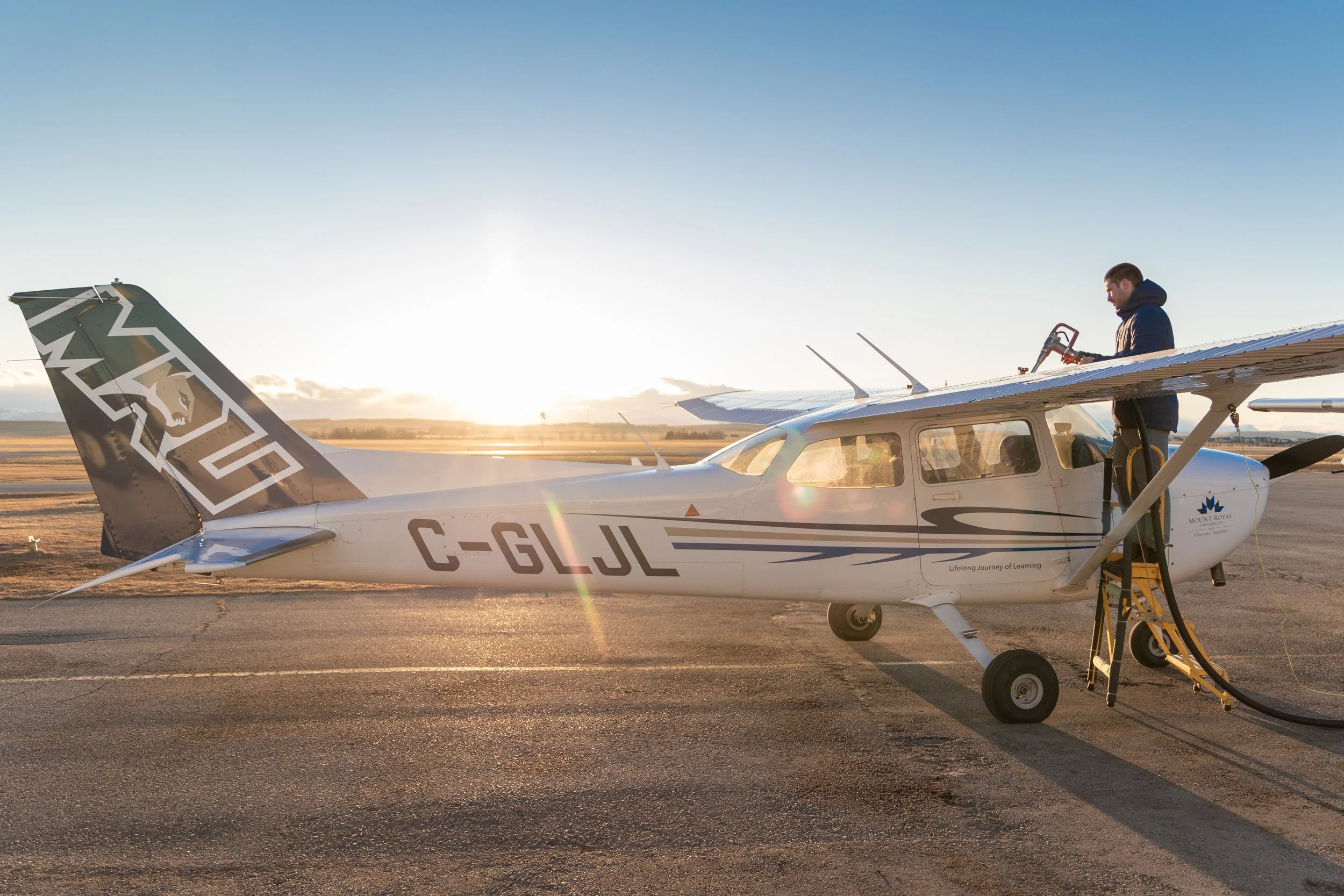 A small airplane on the tarmac with a man standing on a ladder, fueling or inspecting it as the sun sets in the background.