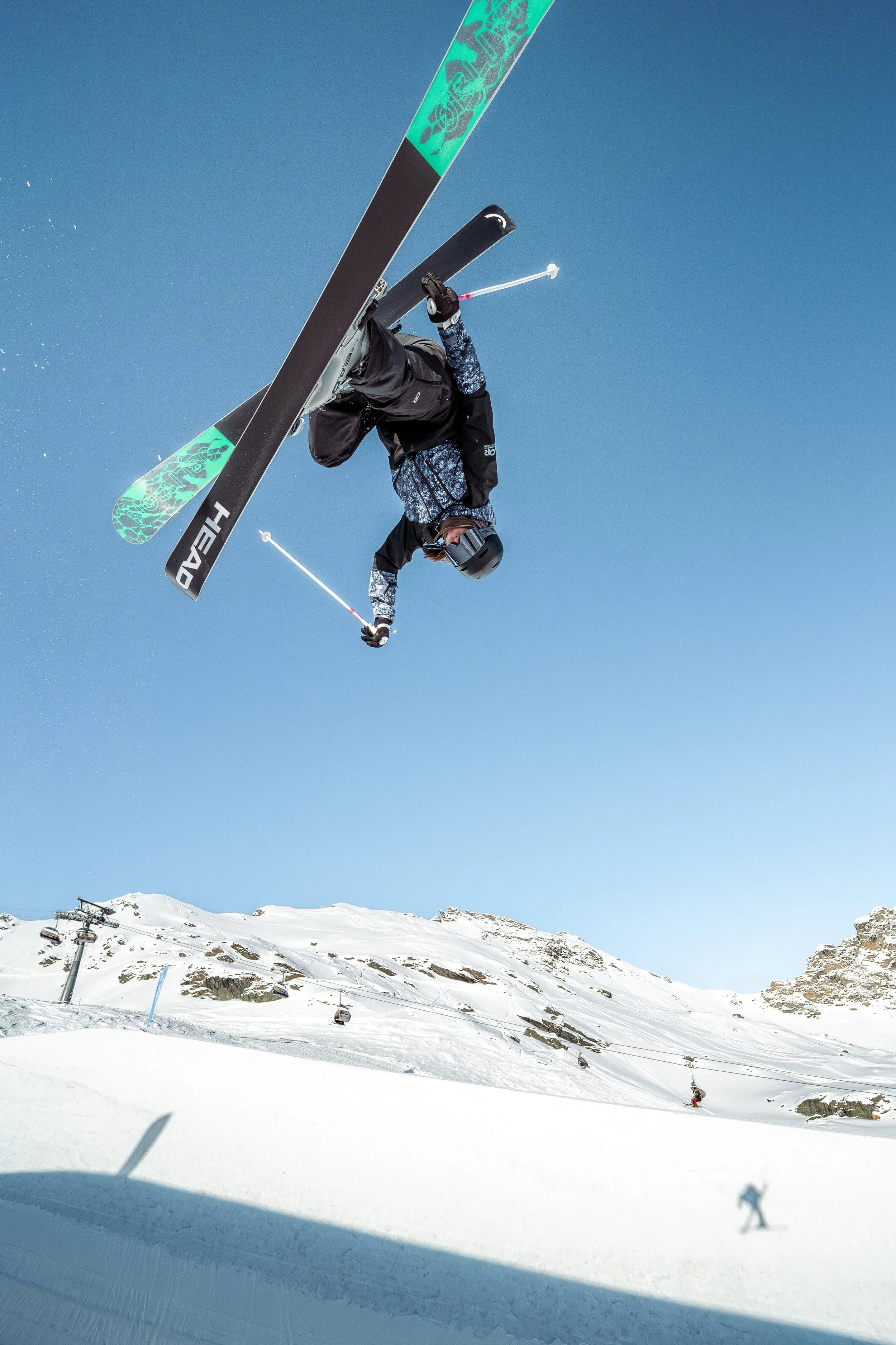 A skier flying through the air with snow and ski lifts visible below and scenic mountains in the background under a clear blue sky.