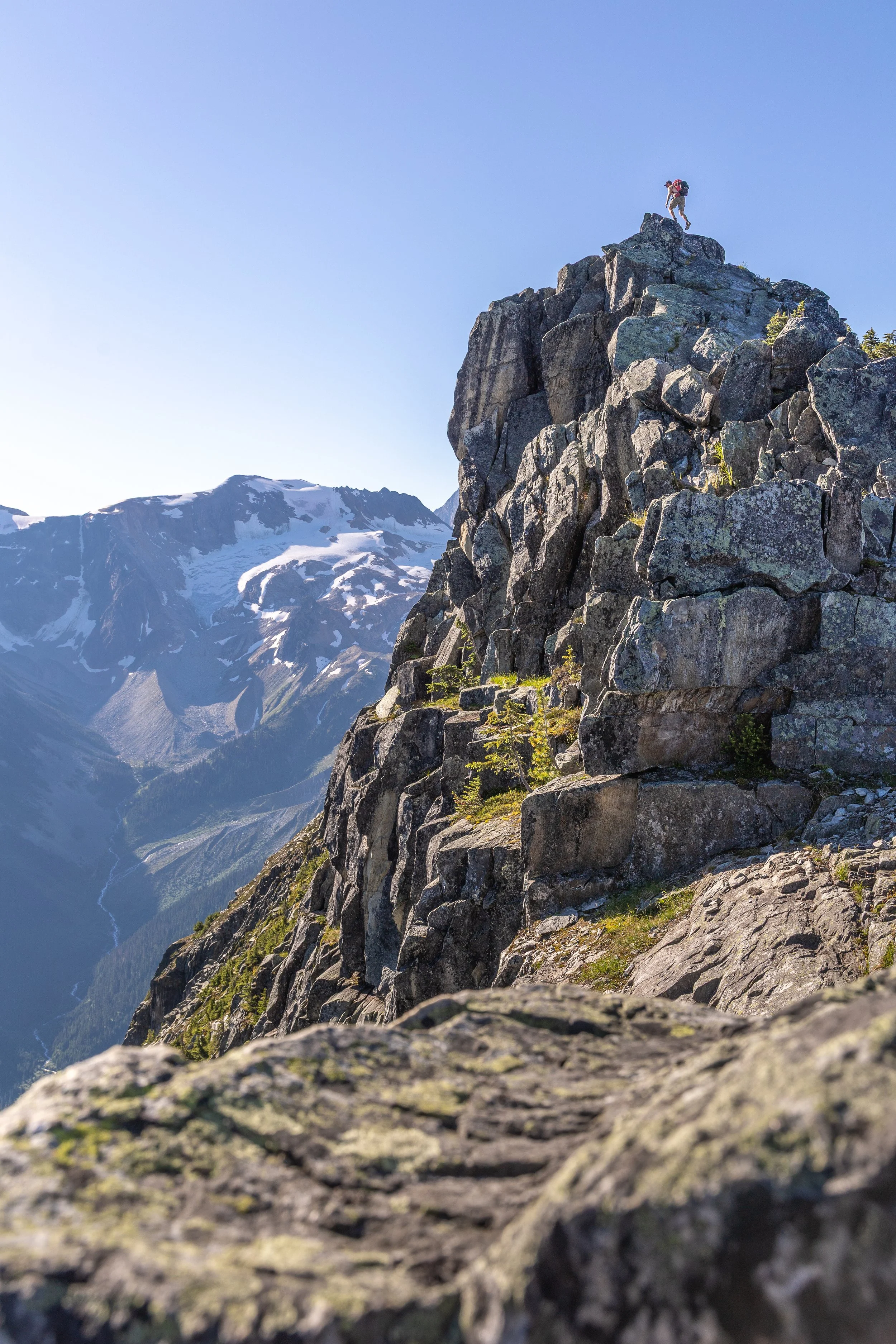A person with a backpack climbing on rugged rocks on a mountain, snow-capped peaks visible in the background under a clear blue sky.