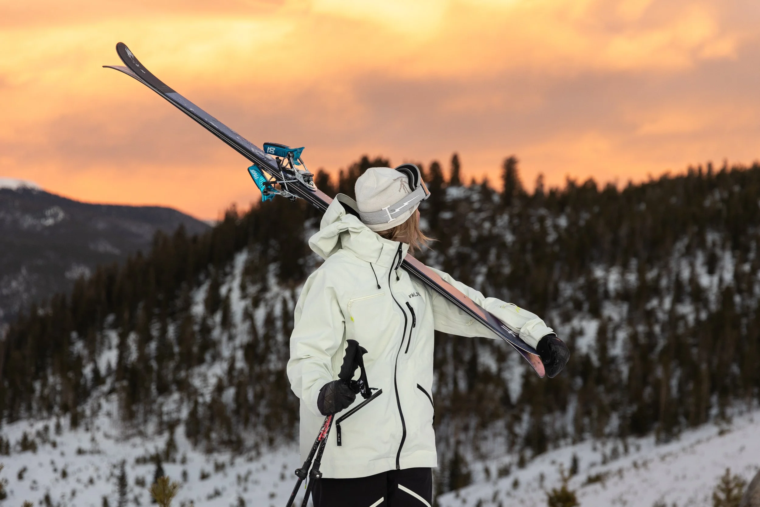 Person in winter gear carrying skis over shoulder on snowy mountain during sunset