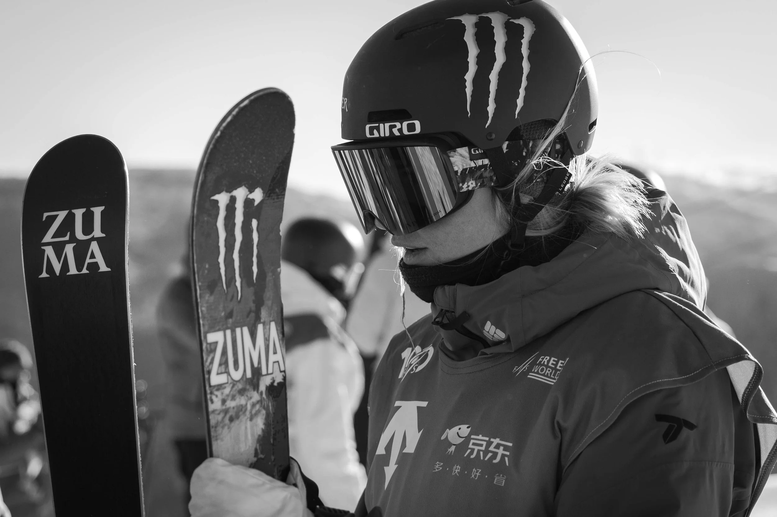 A woman in ski gear and a helmet looks at her skis, with other skiers in the background on a snowy mountain.