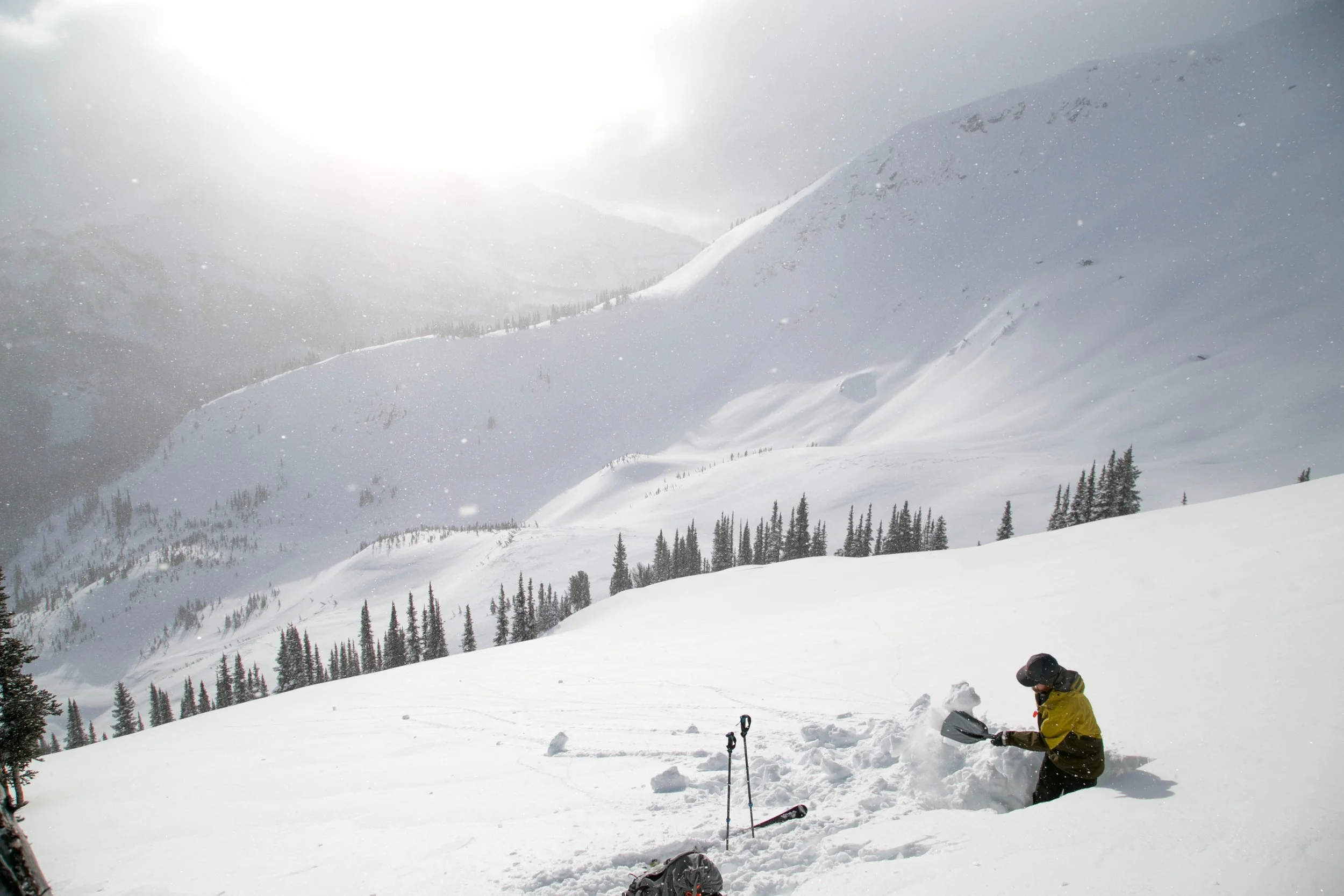 A person in a yellow jacket and black helmet shoveling snow on a snowy mountain landscape with sparse trees and distant snow-covered peaks, under cloudy skies with the sun peeking through.