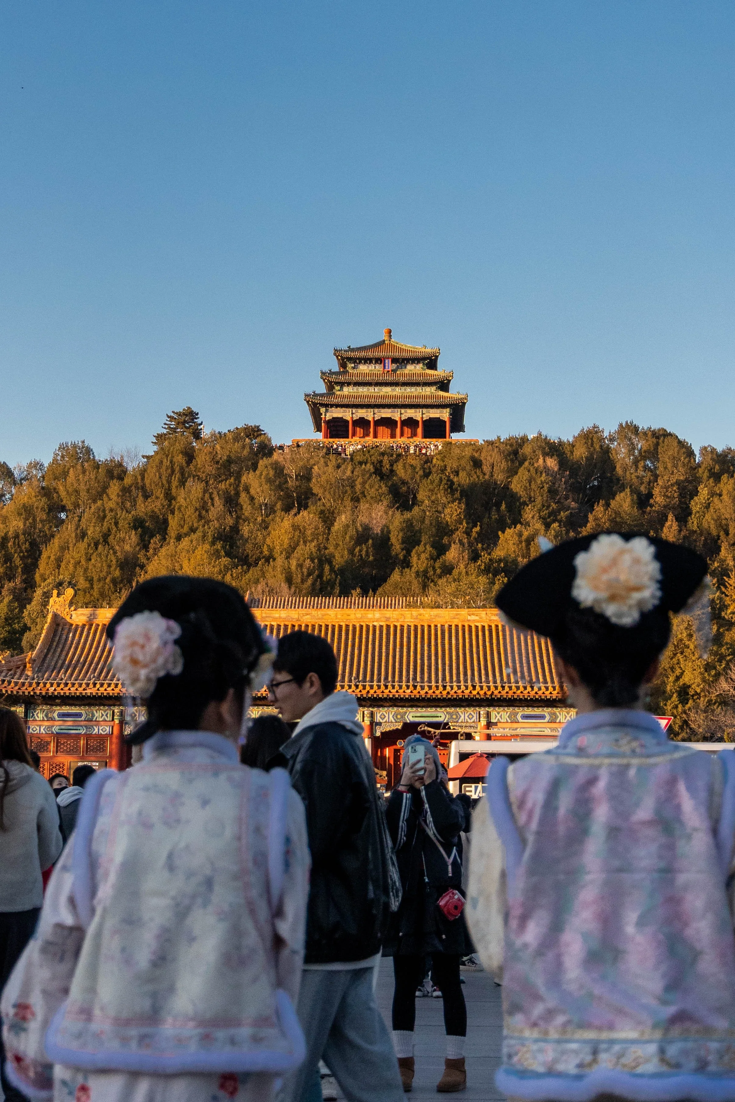 Tourists, including two women dressed in traditional clothing, in front of a historic Chinese building on a hill with trees, under a clear blue sky.