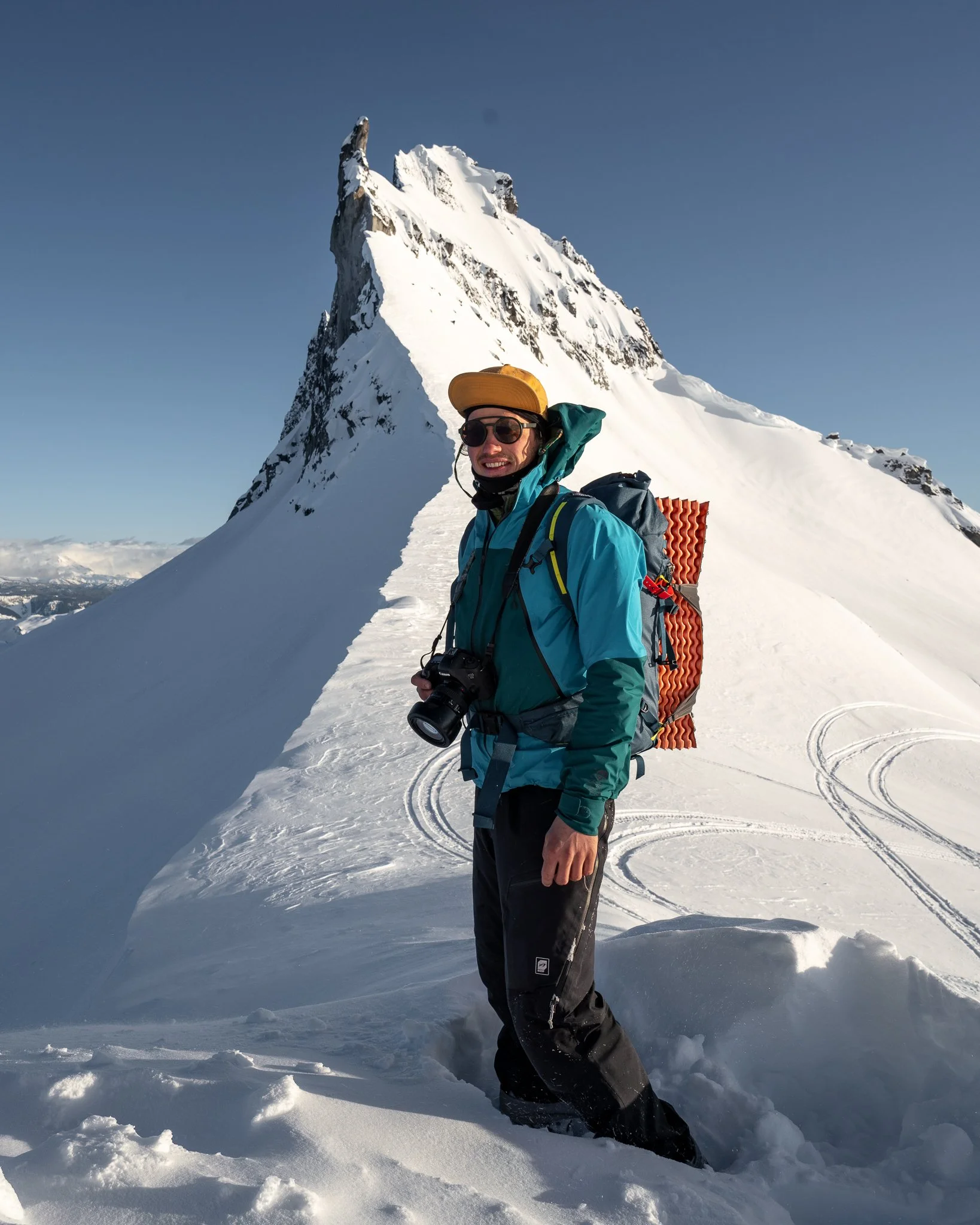 A man in outdoor winter gear standing on a snow-covered mountain path with a mountain peak in the background, holding a camera.