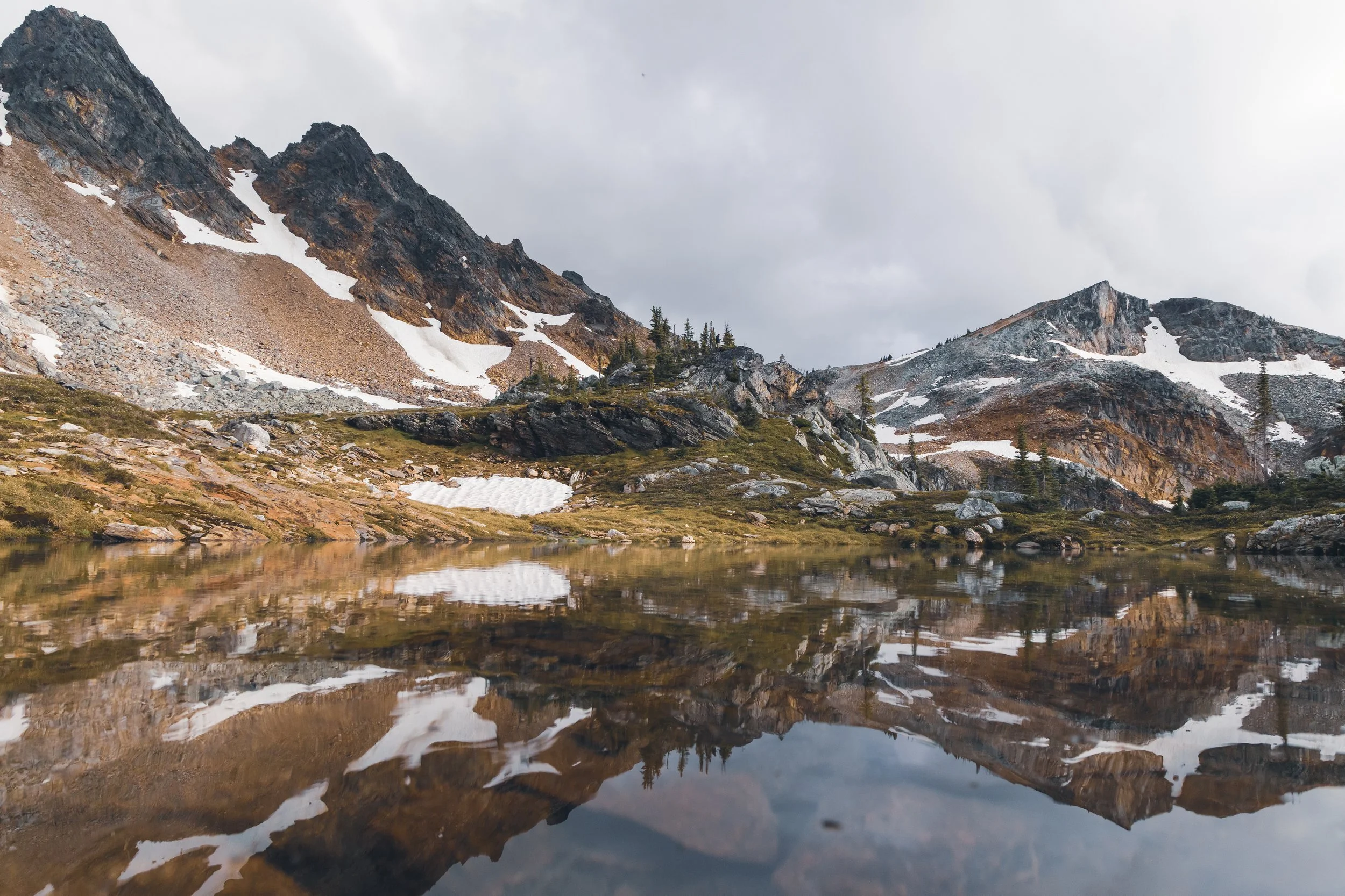 Mountains with patches of snow, rocky slopes, and pine trees reflected in a calm lake under a cloudy sky.