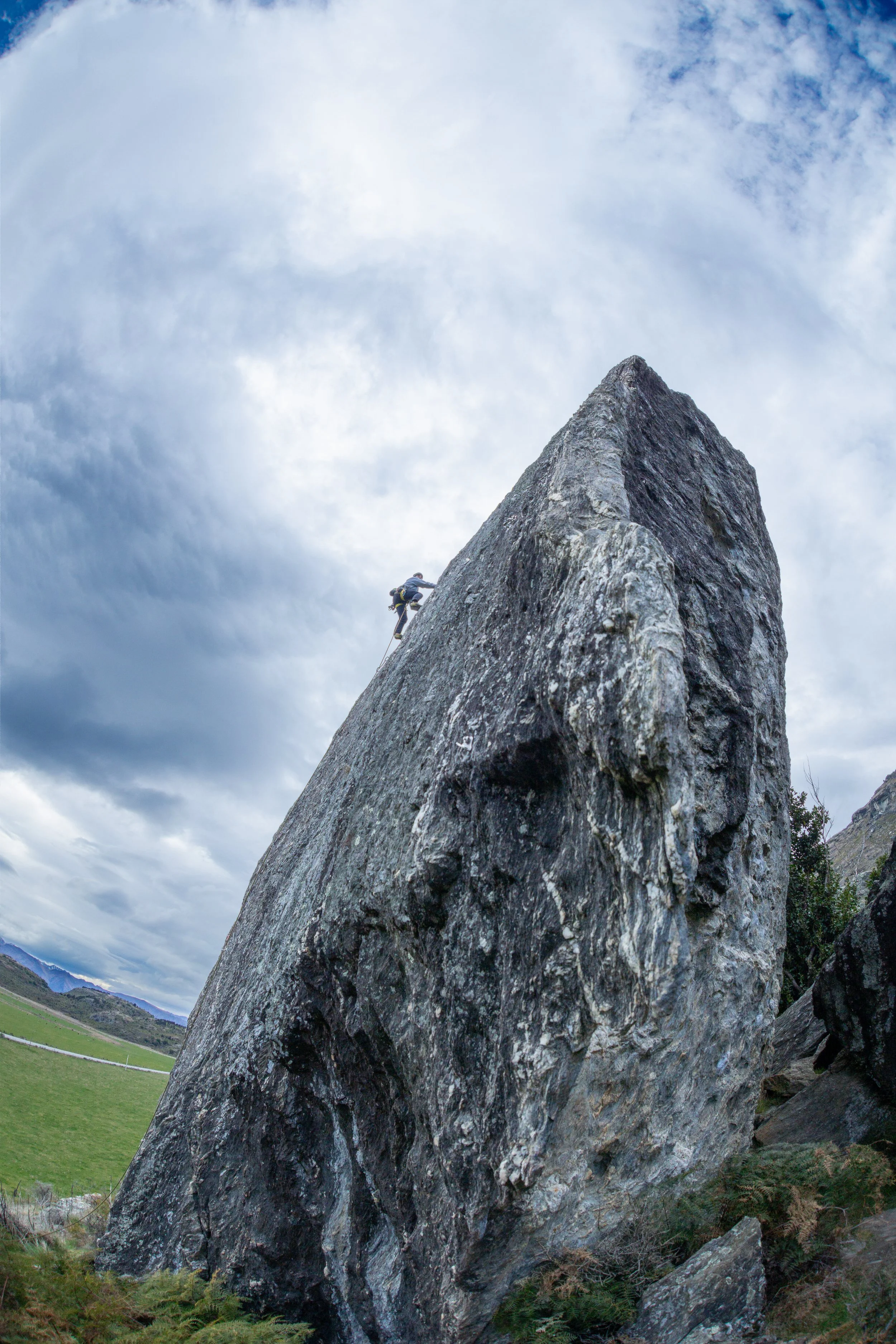 A person climbers a steep, large gray rock formation in a grassy field under a cloudy sky.