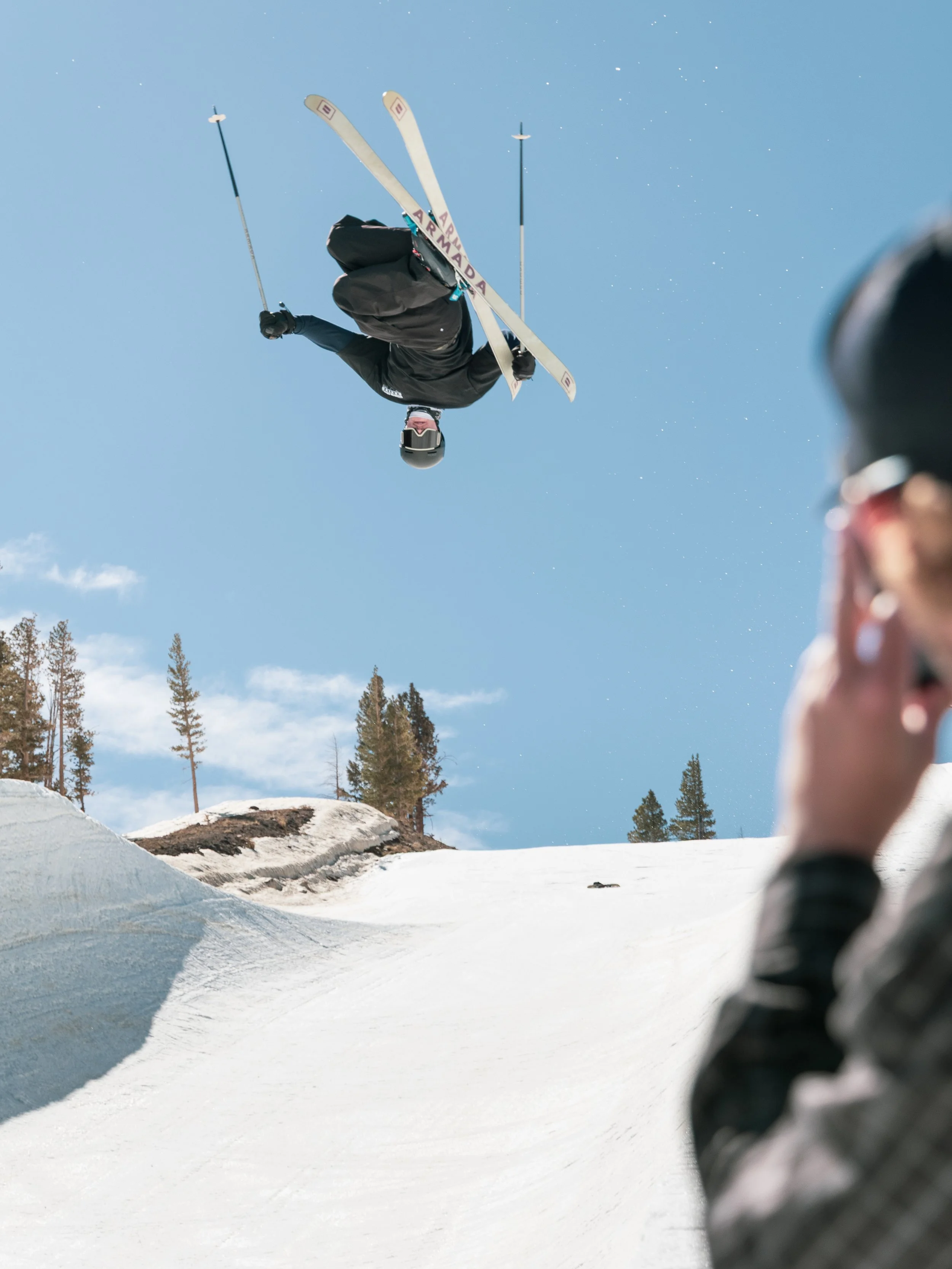 A skier performing a jump in the air on a snowy slope, with trees and a clear blue sky in the background. A person in the foreground is watching and recording the scene.
