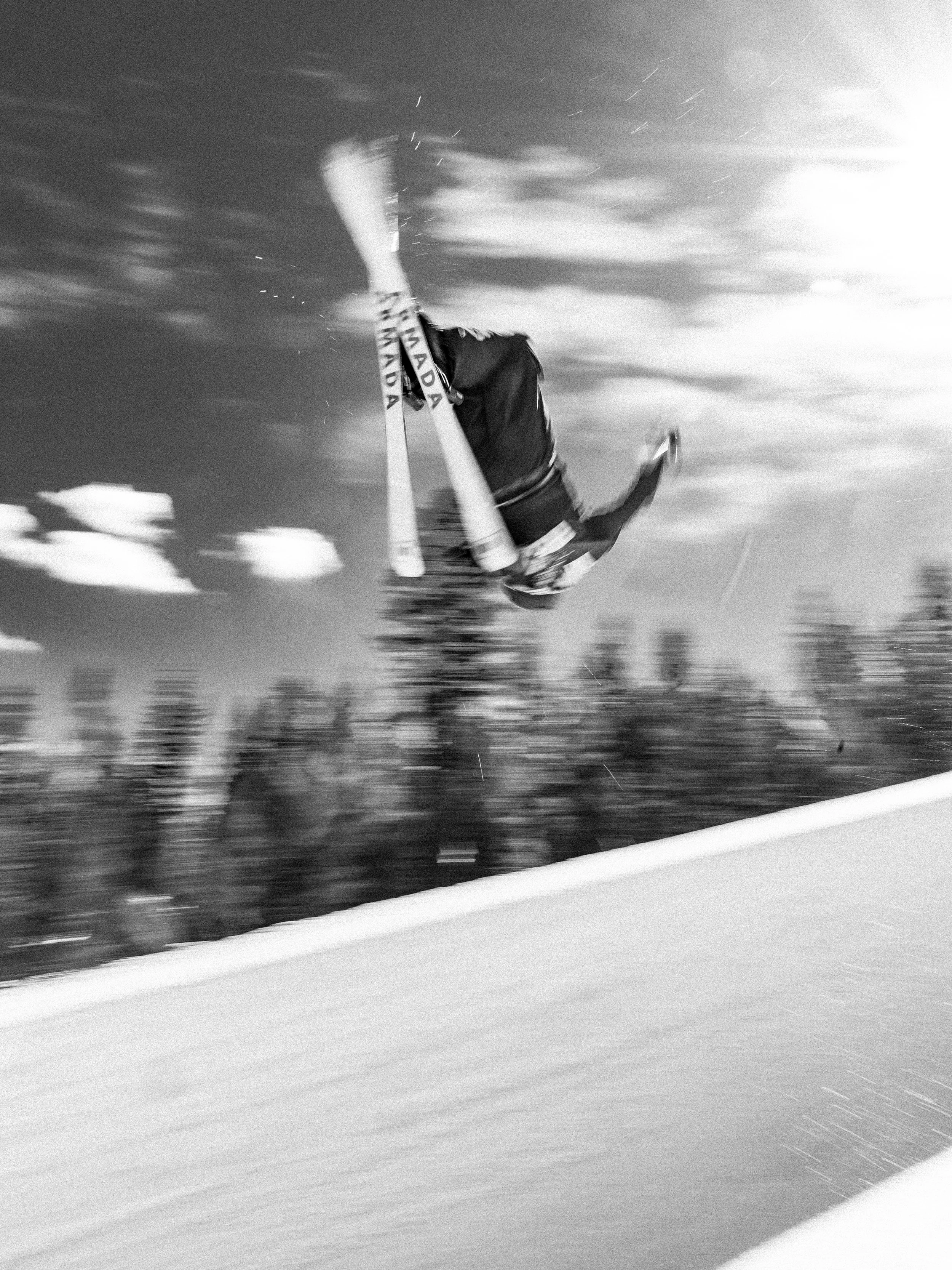 A skier in mid-air performing a trick during a jump on a snowy slope, with a blurred background of trees and clouds.