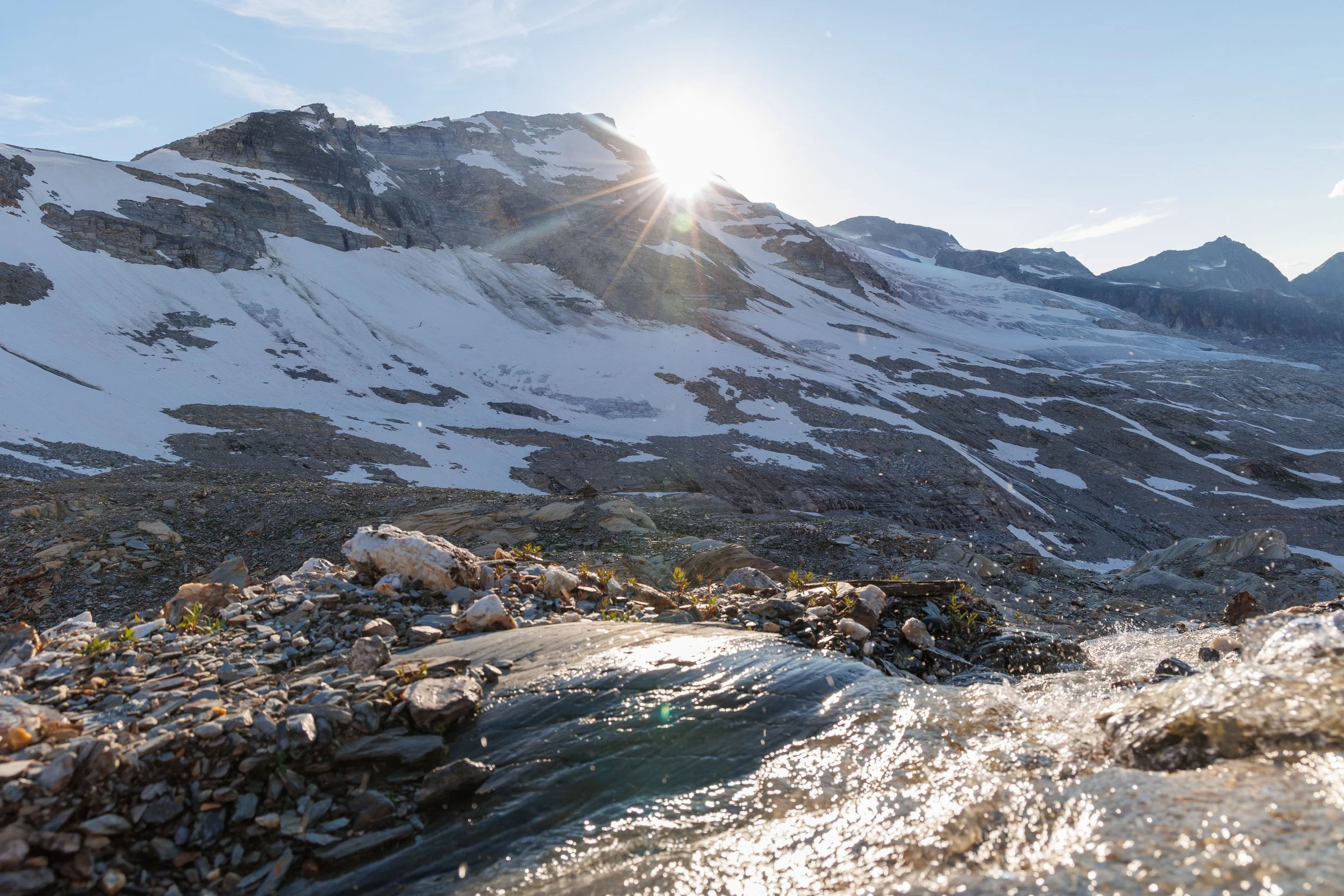 Snow-covered mountain landscape with a rocky and gravelly foreground, a flowing stream, and sunlight peeking over the mountain peaks.
